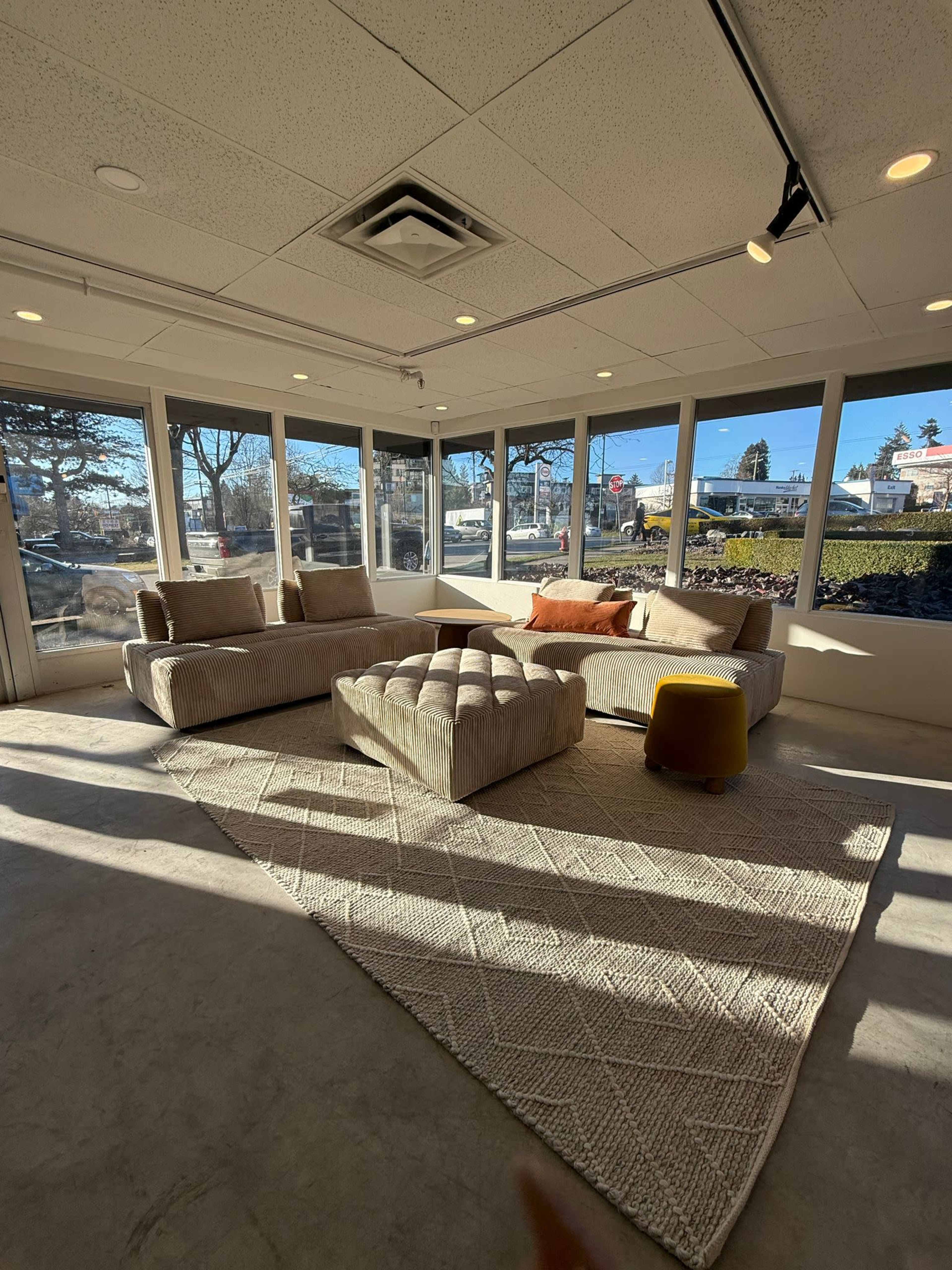 A modern living space features a sectional sofa, a tufted coffee table, and a yellow stool on a textured area rug, all set within a room surrounded by large windows.