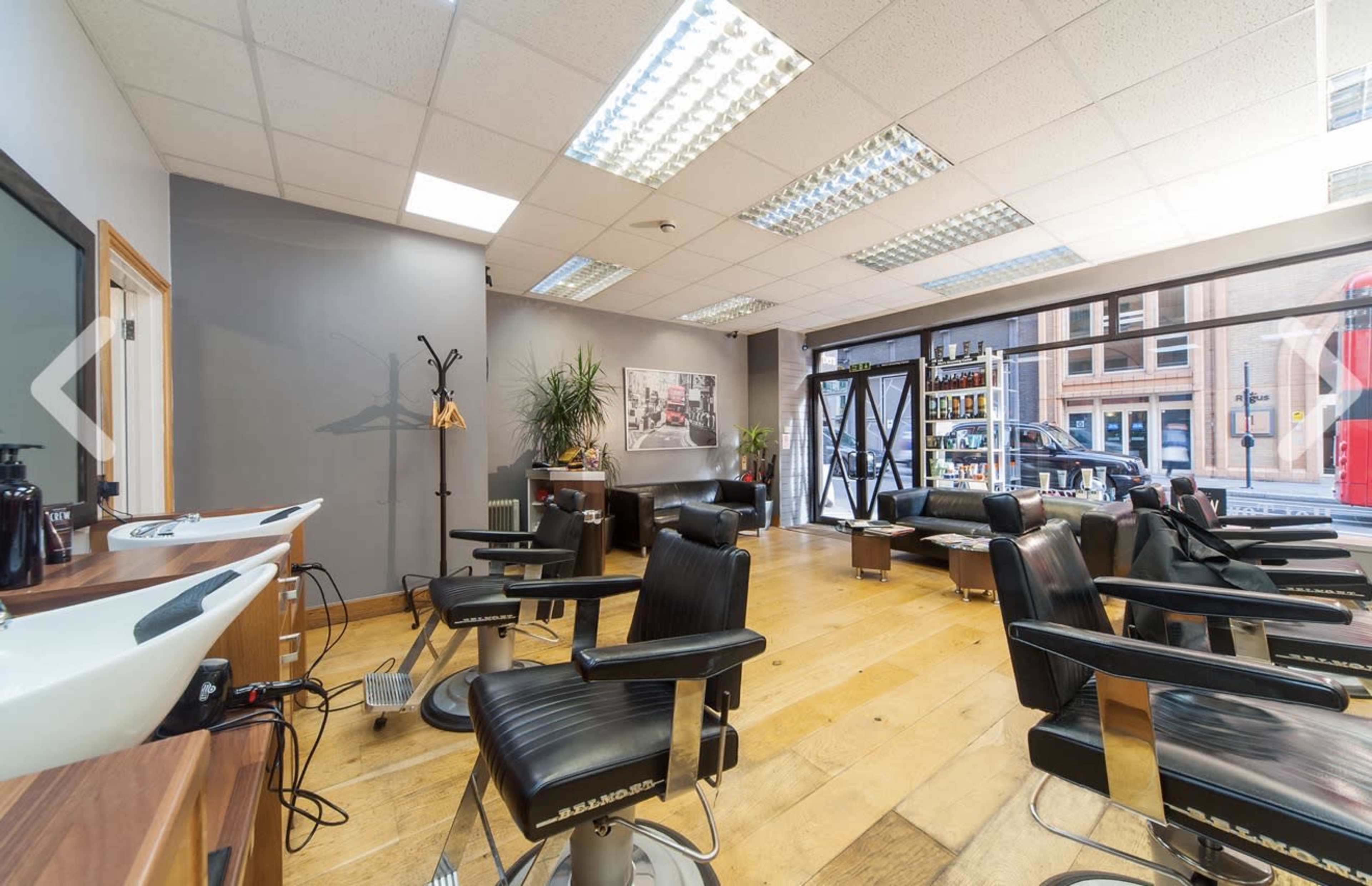The image shows a modern barber shop interior with several barber chairs arranged around a wooden floor and large windows letting in natural light.