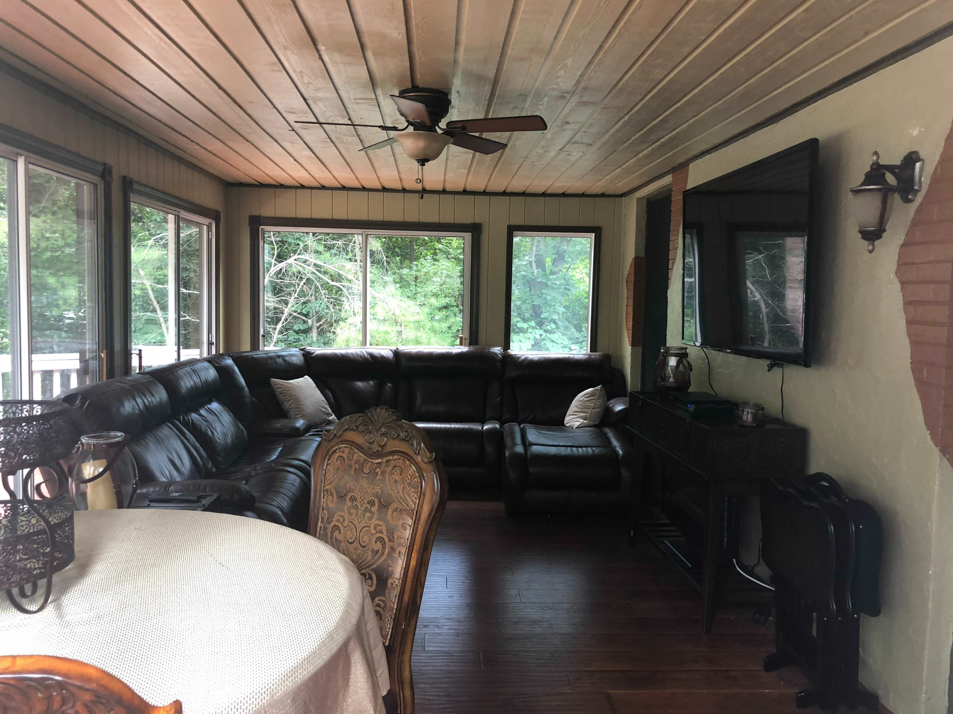 A screened-in porch furnished with a dark leather sectional sofa, a round dining table, and a television mounted on the wall, surrounded by large windows that offer a view of greenery outside.