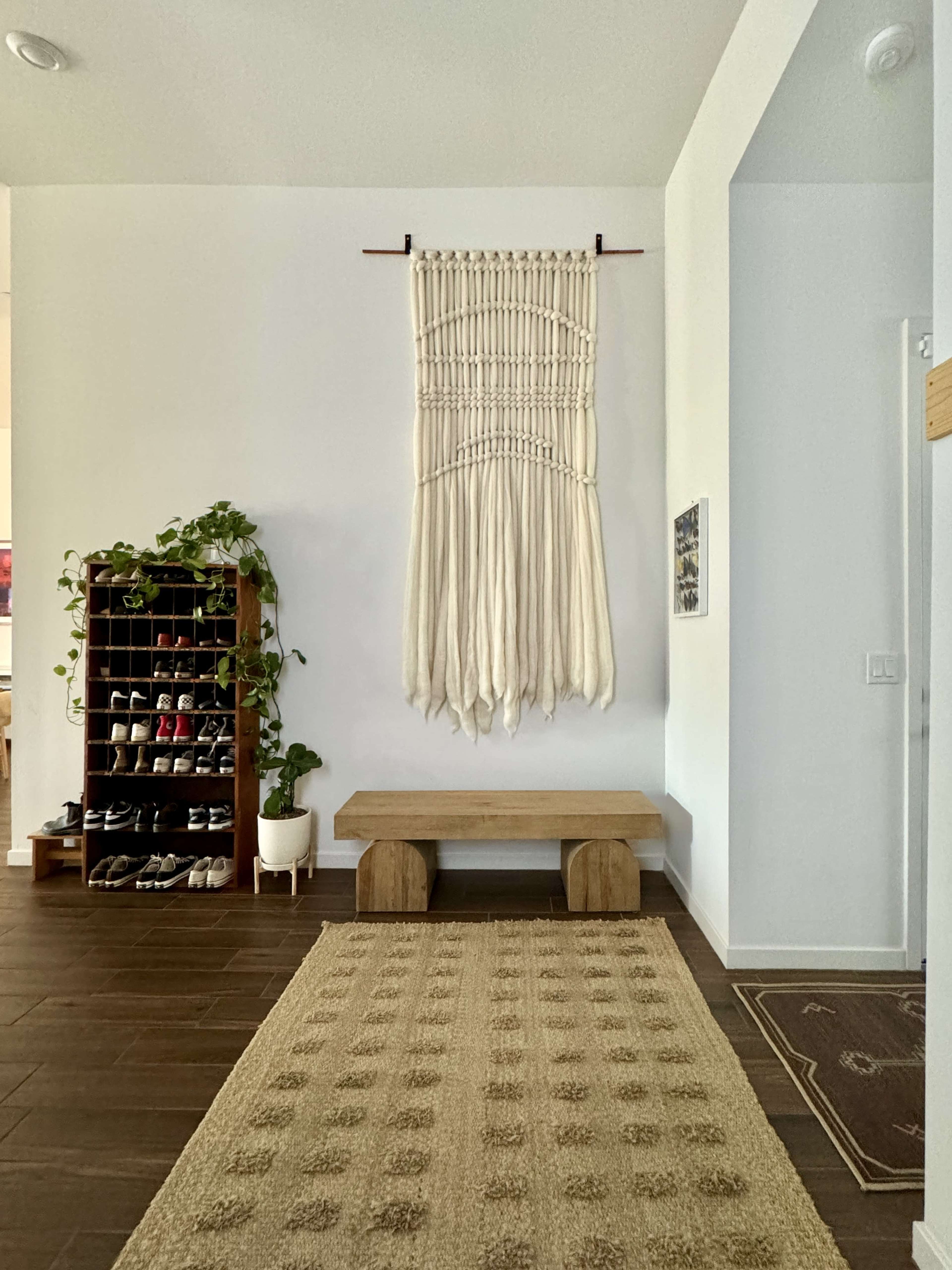 The image shows a home entryway with a woven wall hanging, a wooden bench, a shoe rack filled with shoes, and a textured rug on the floor.