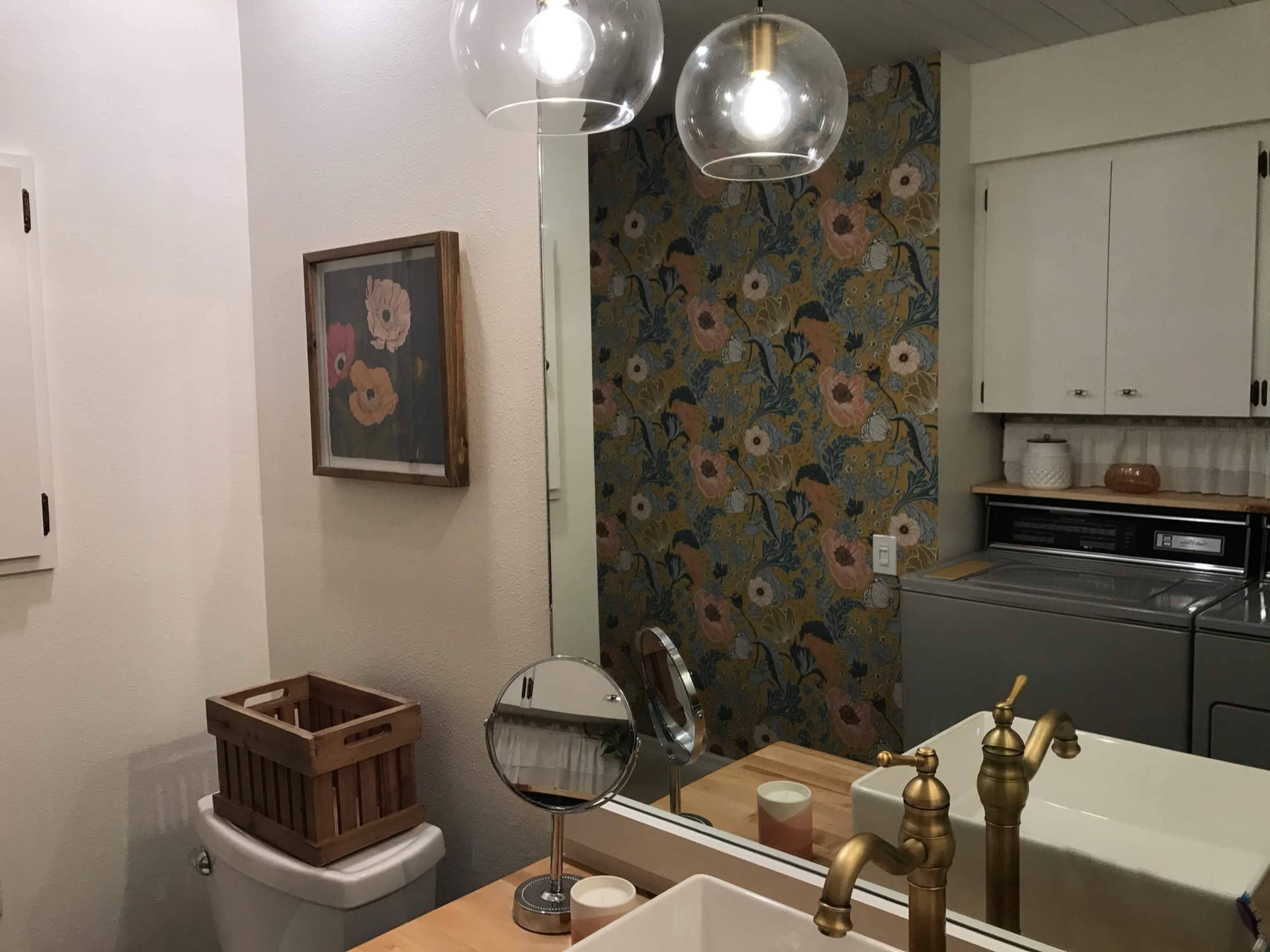 A bathroom features a wall with floral wallpaper, a large mirror, a sink with a gold faucet, a laundry machine, and a wooden basket for storage.