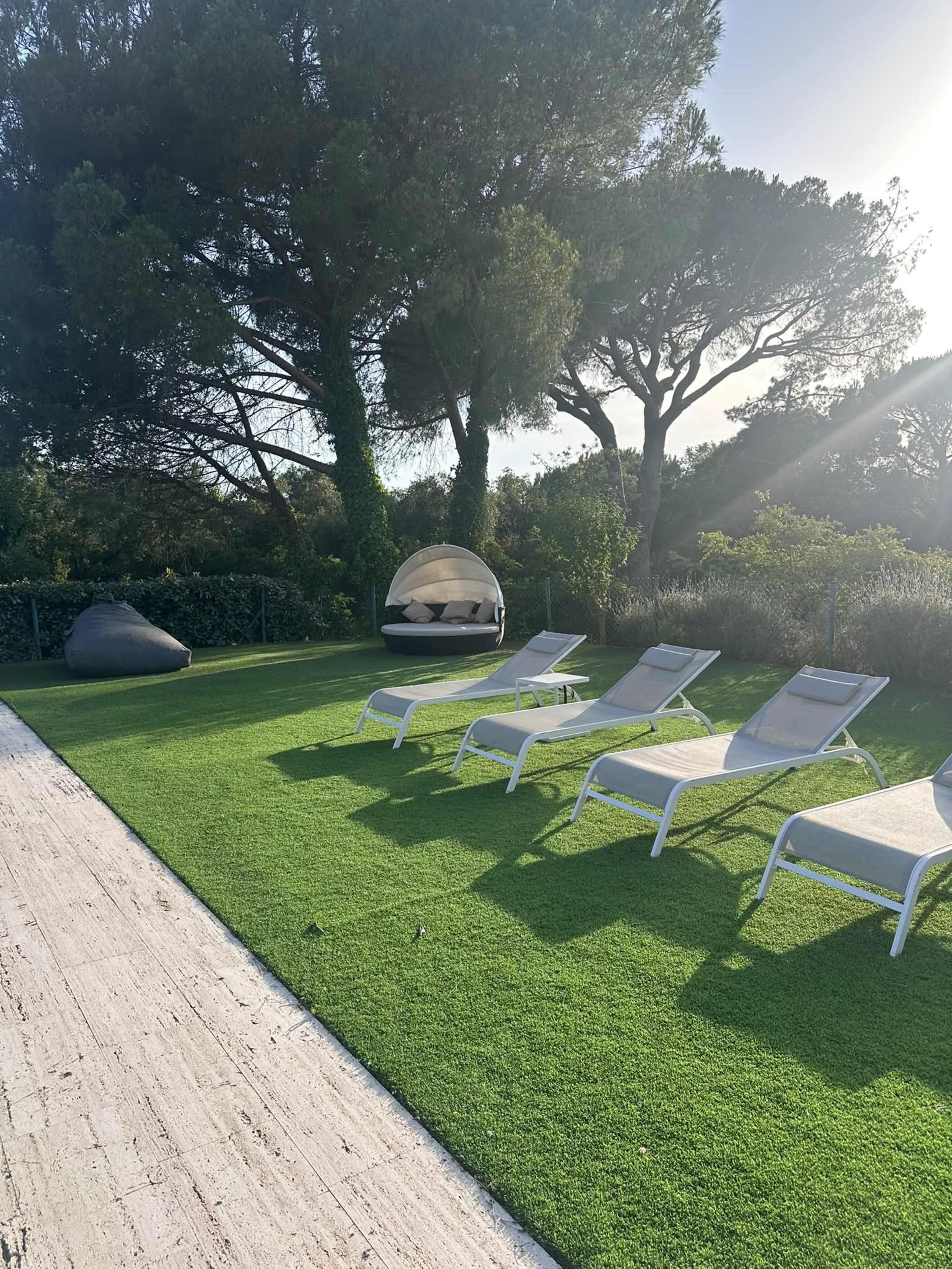 The image shows a landscaped outdoor area featuring several lounge chairs on artificial grass, a round shaded seating pod, and trees in the background.