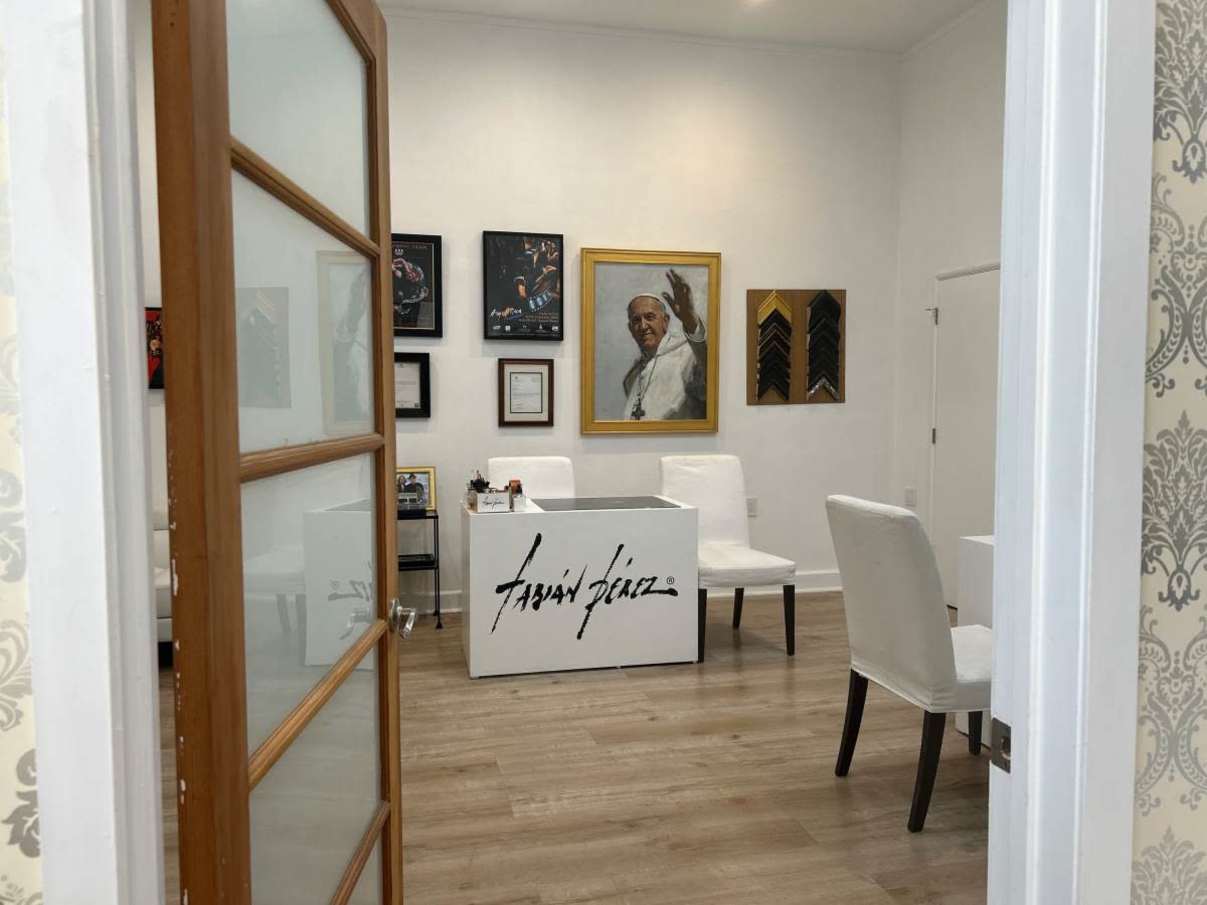 The image shows an interior view of an office or gallery featuring a table with the name "Fabian Pérez," artwork on the walls, and white chairs.