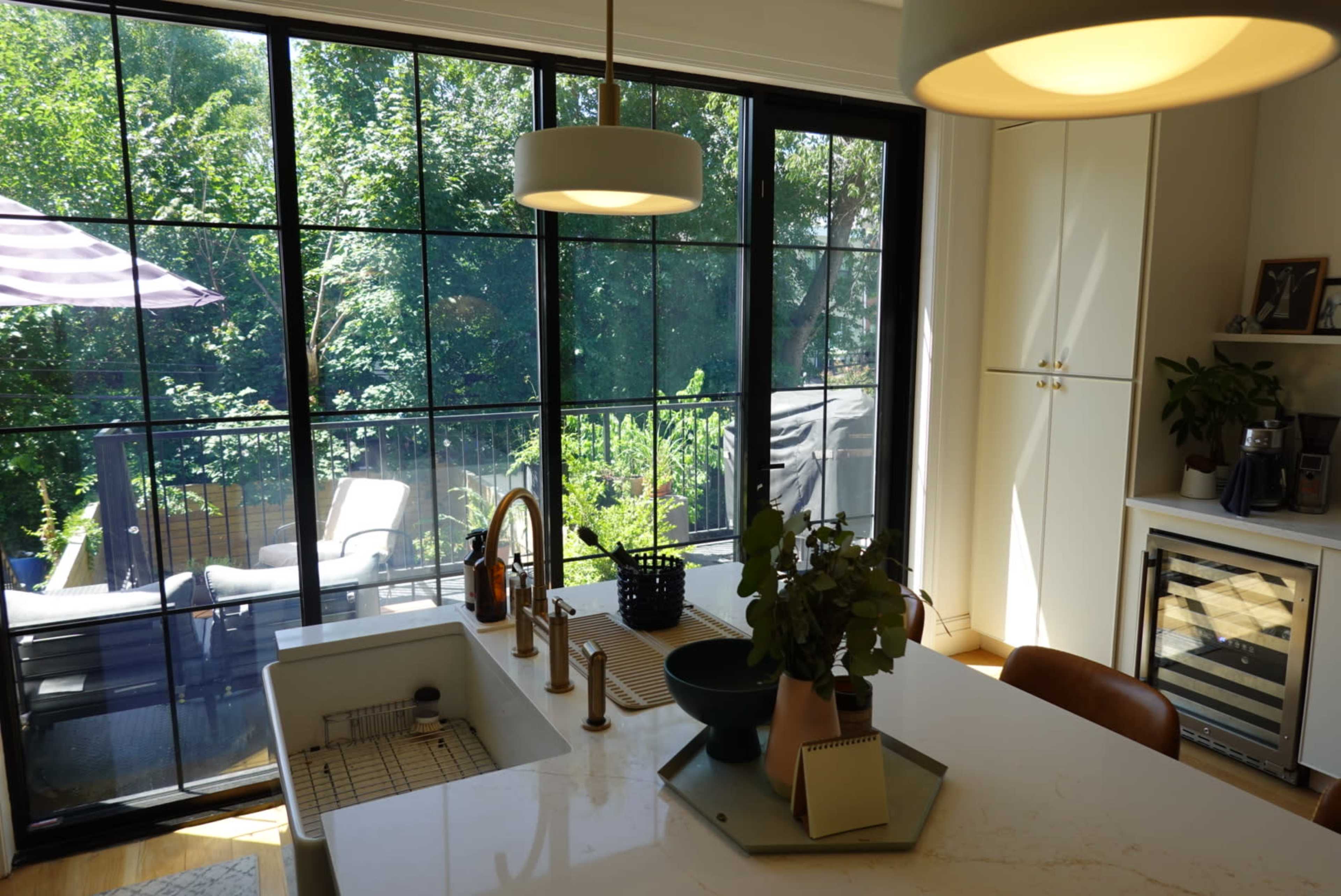 The image shows a modern kitchen with large windows overlooking a green outdoor space, featuring a central island and stylish fixtures.