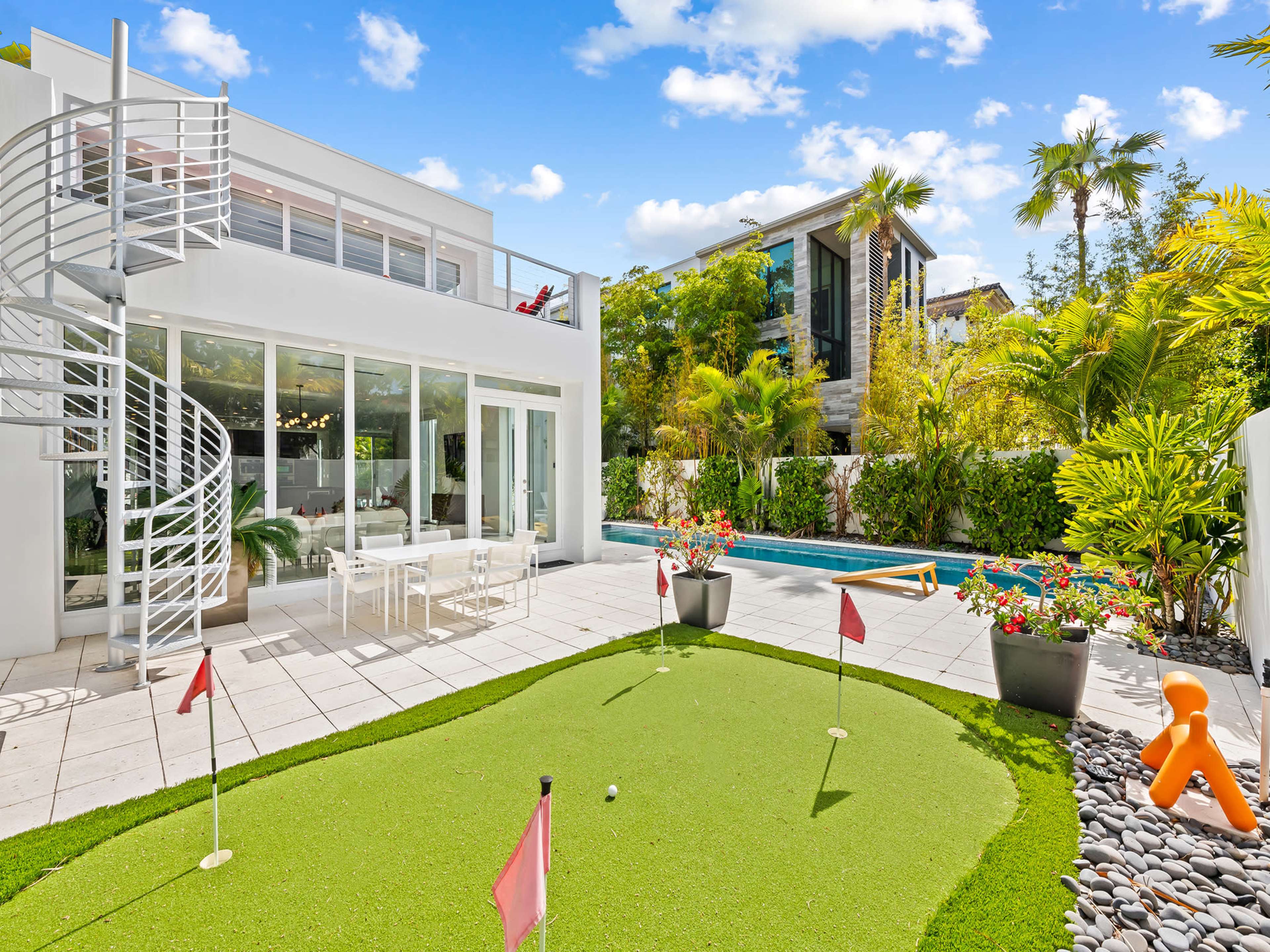 The image shows a modern home with a spiral staircase, a mini-golf area, a swimming pool, and lush greenery surrounding the backyard.
