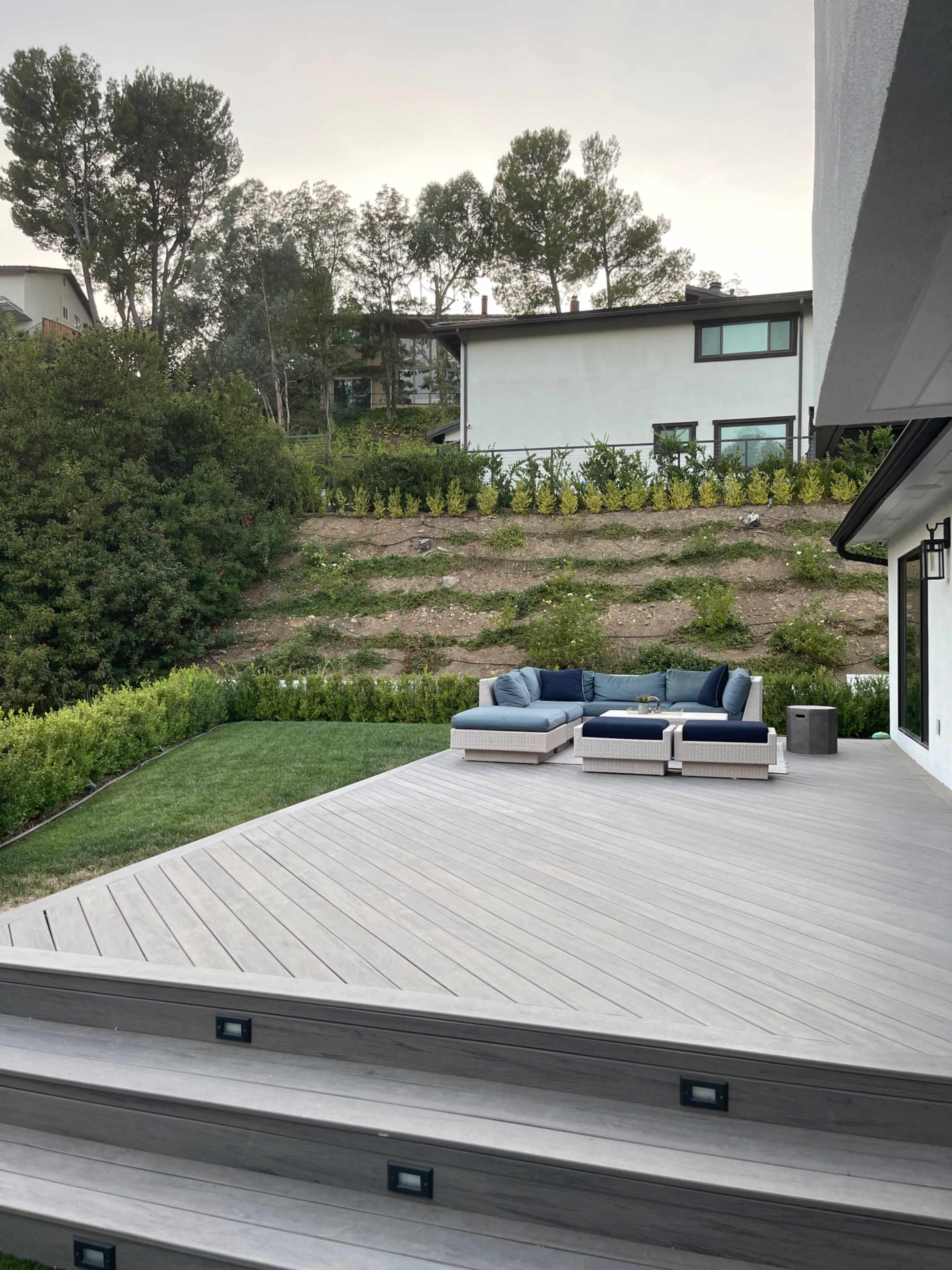 A wooden deck with outdoor seating on a lawn, surrounded by greenery and a hillside.
