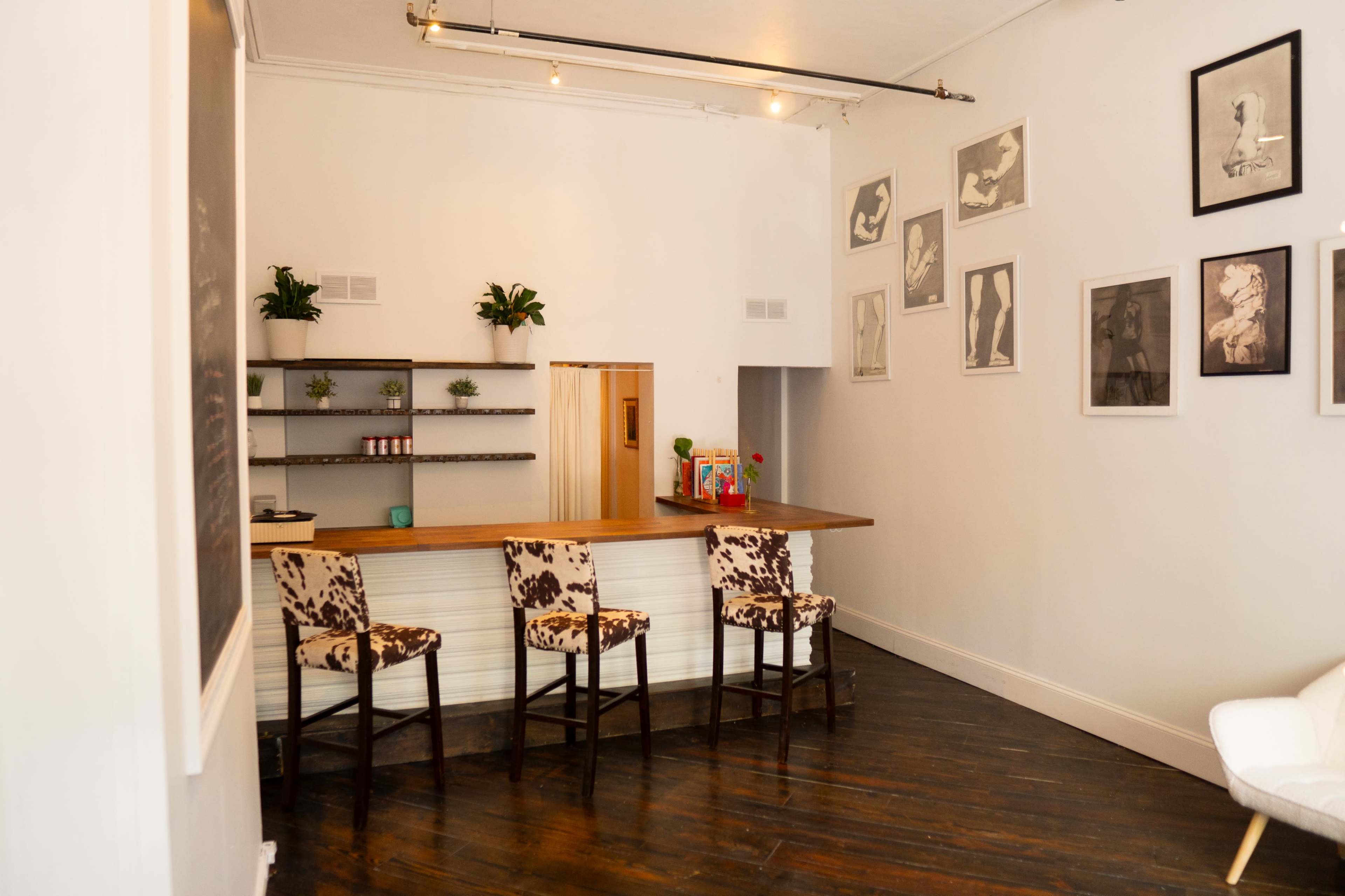 A minimalist interior with a small reception area featuring a wooden counter, three patterned stools, and framed artwork on the walls.