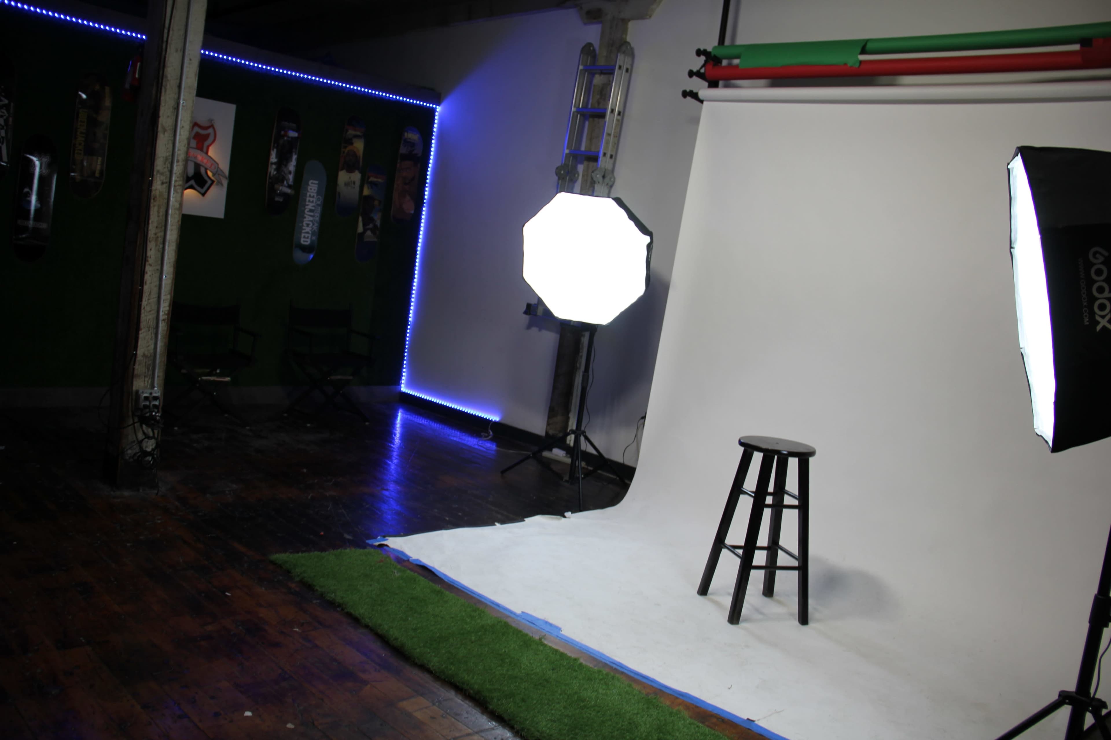 A photography studio with a white backdrop, a stool, and lighting equipment, complemented by green turf on the ground and neon blue accents along the walls.