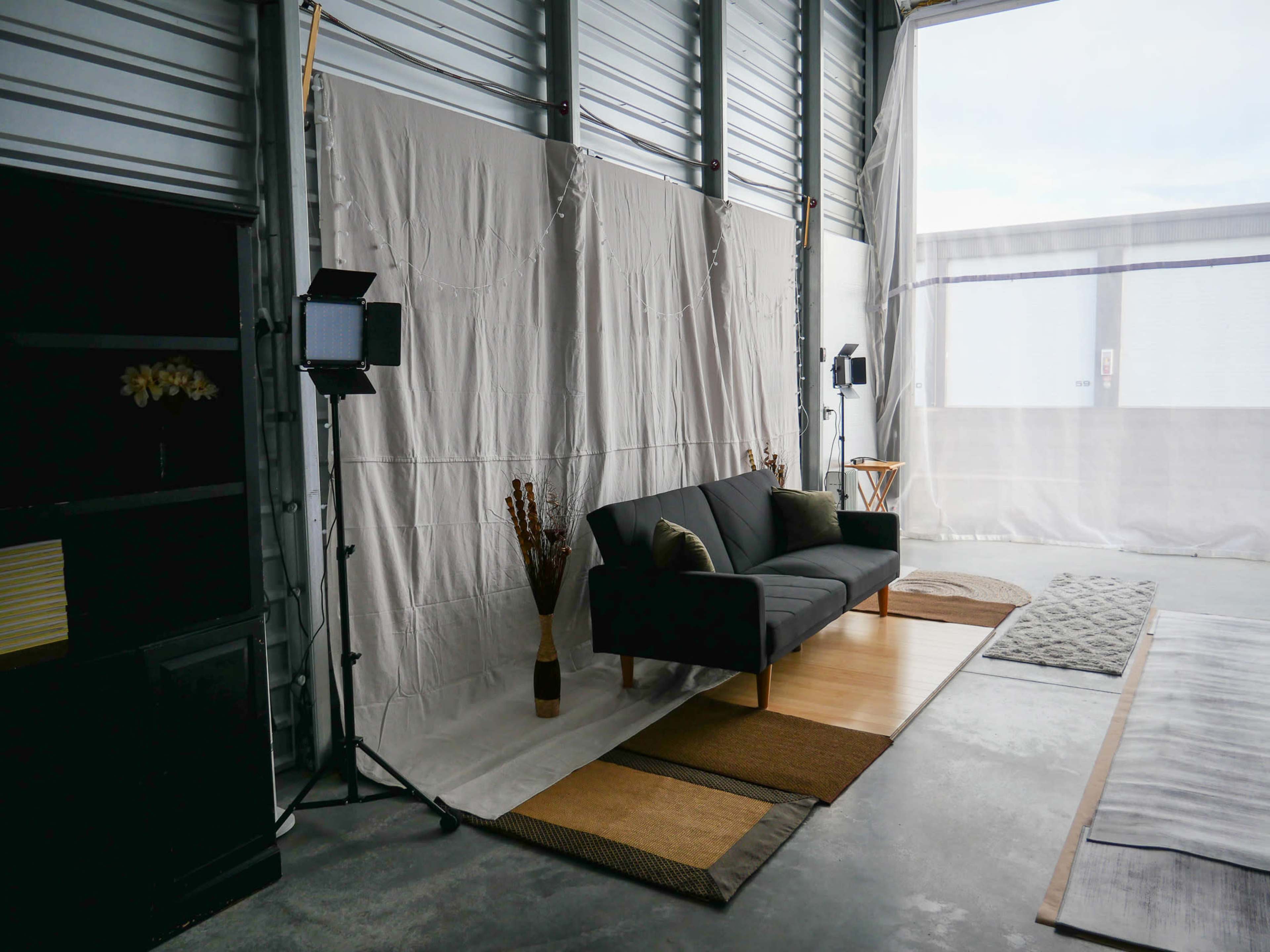 A minimalist interior with a black sofa, a small wooden table, and studio lighting set up in a space with white draped walls and concrete flooring.