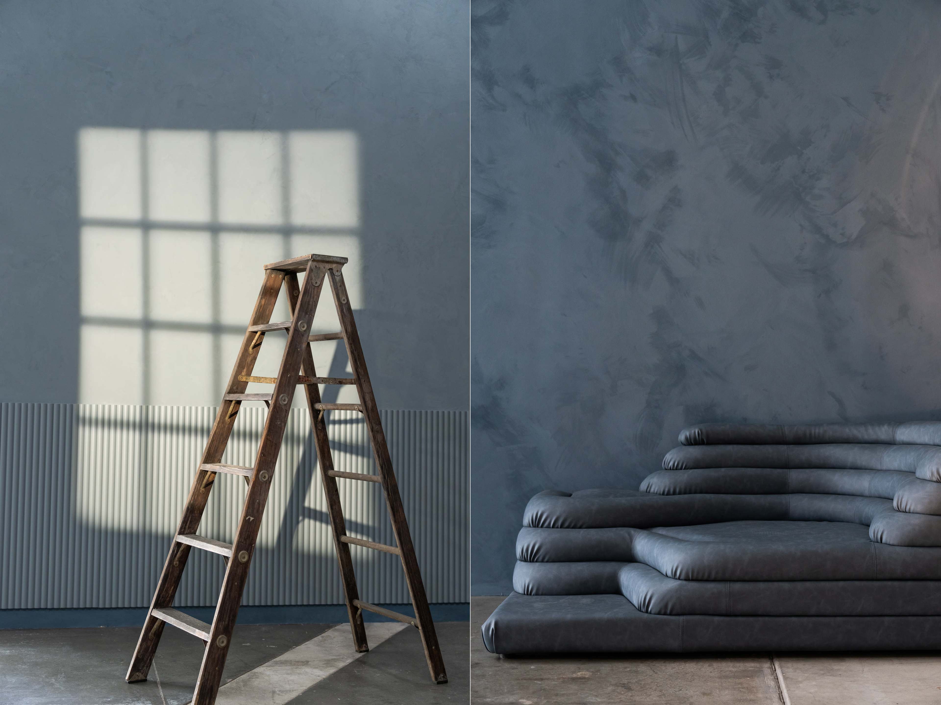 A wooden ladder positioned beside a stack of gray cushions against a textured blue wall with natural light casting a shadow of a window.