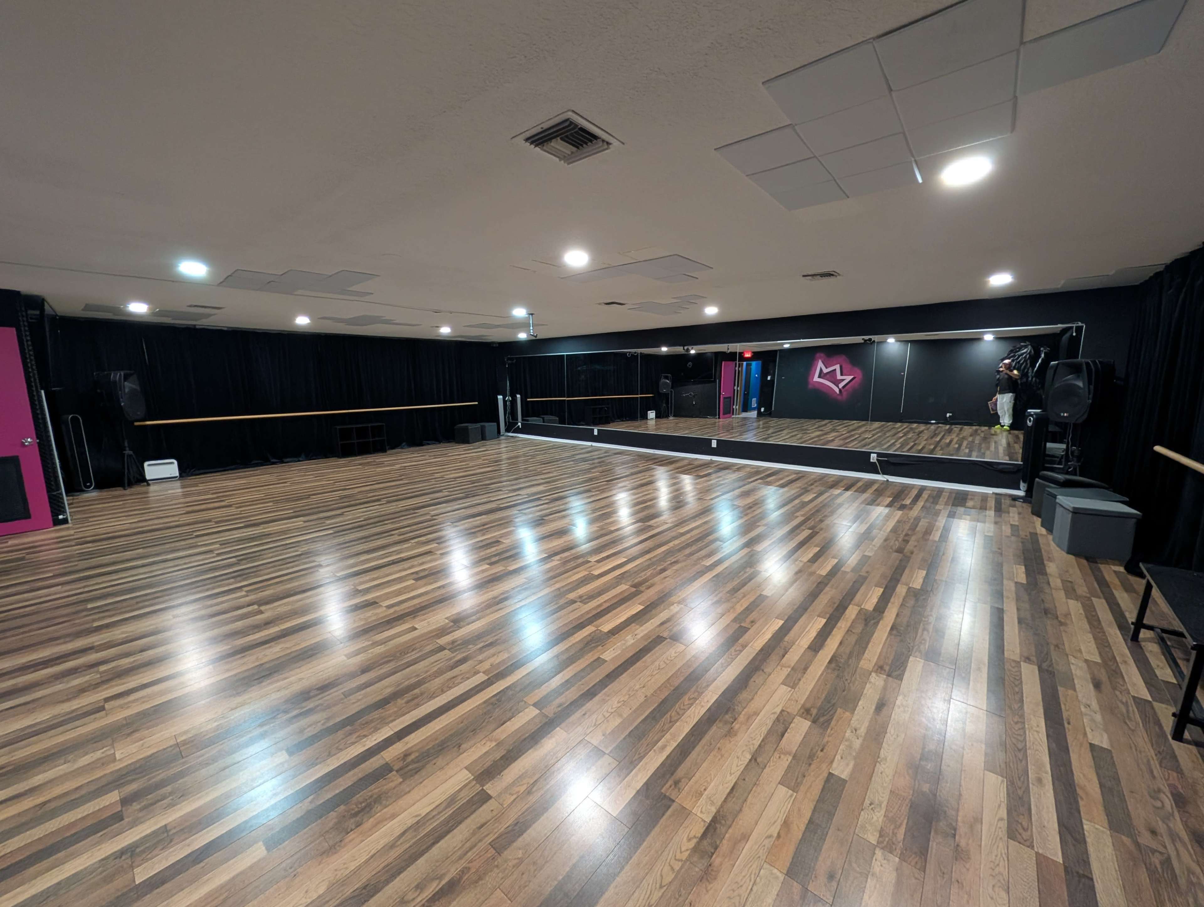 The image shows a large, empty dance studio with a wooden floor, mirrored wall, and black curtains.