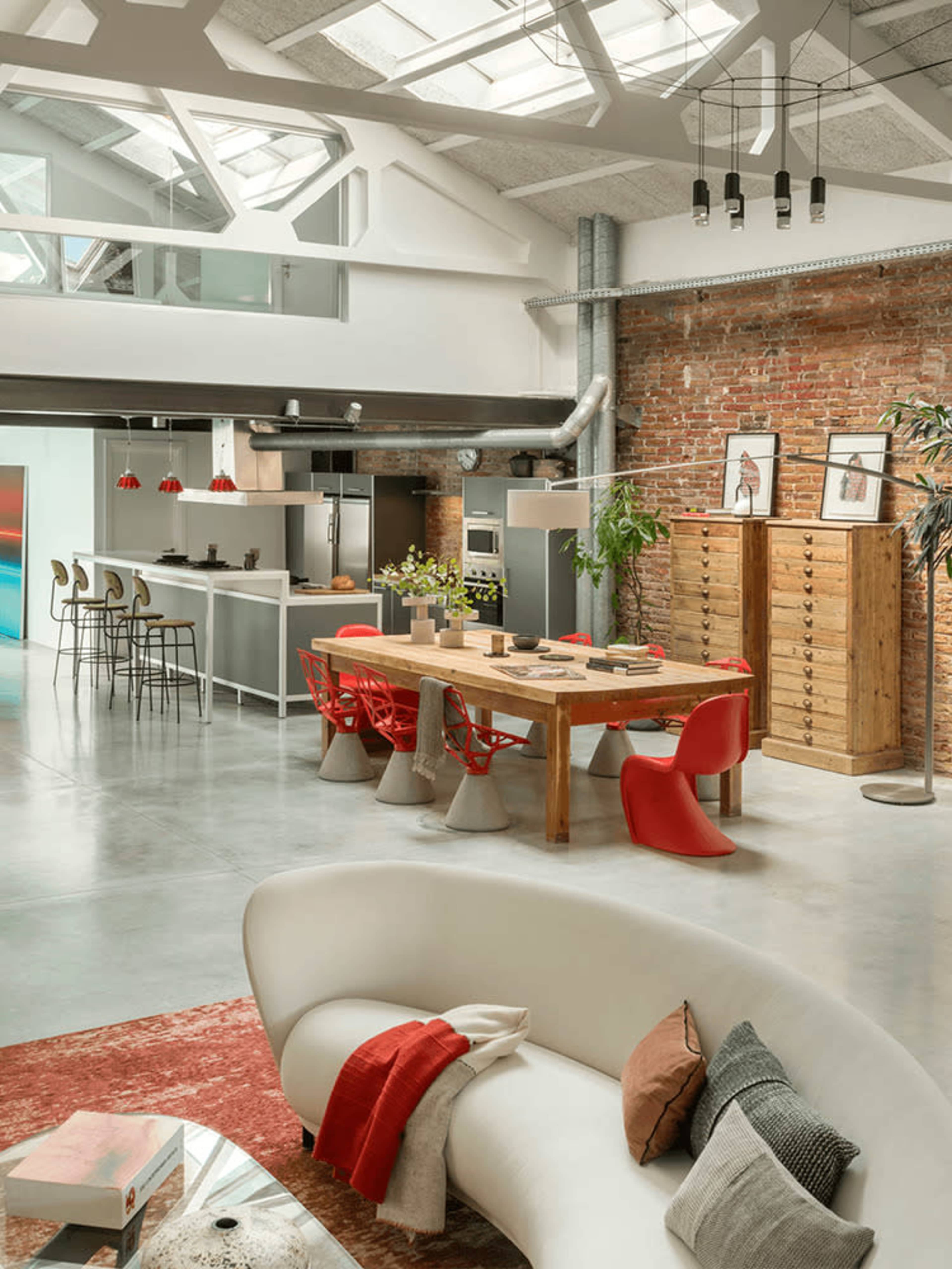The image features a modern open-concept kitchen and dining area with a large wooden table, colorful chairs, and exposed brick walls.