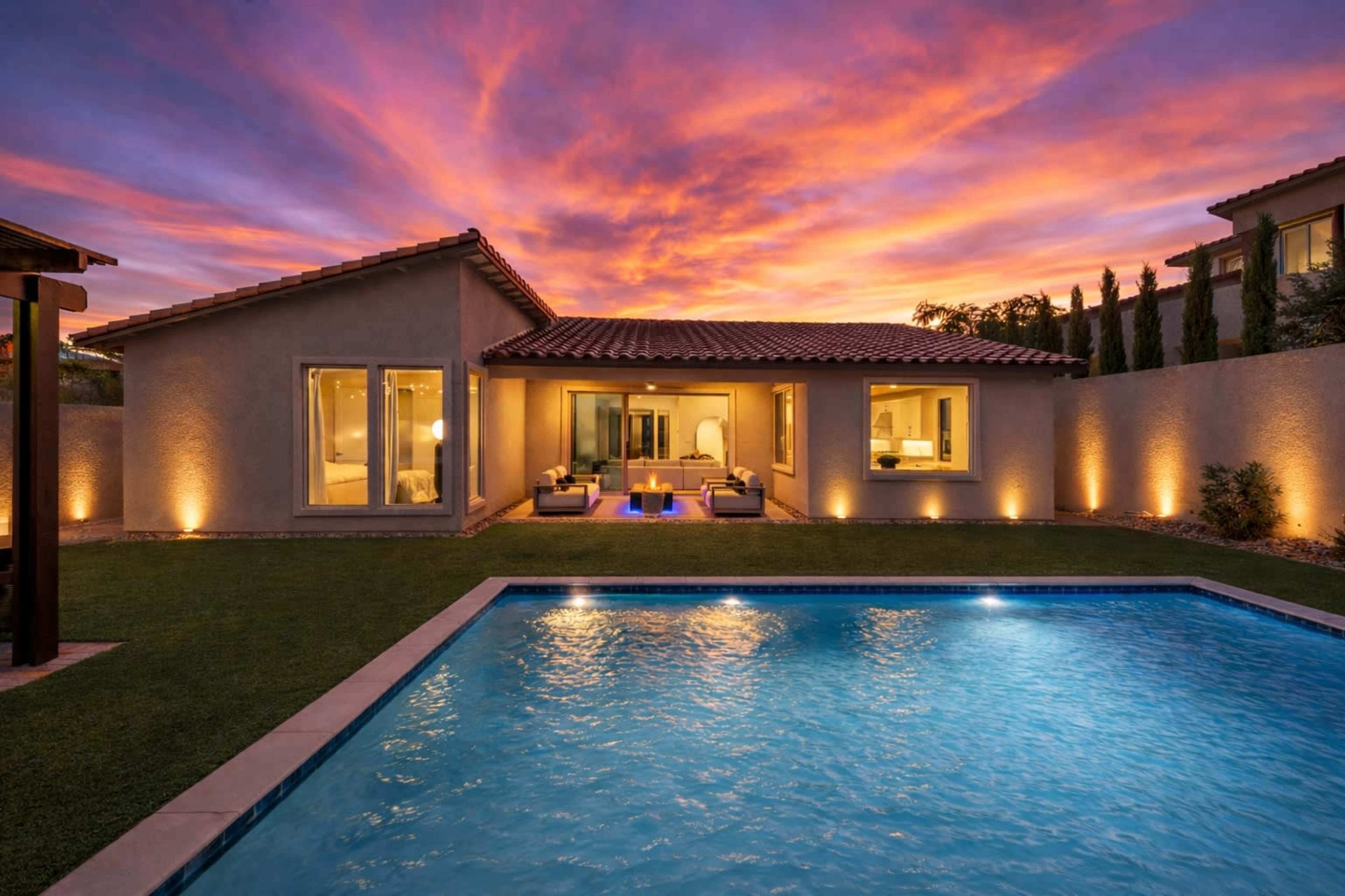 A modern house with large windows and a pool is illuminated by a colorful sunset.