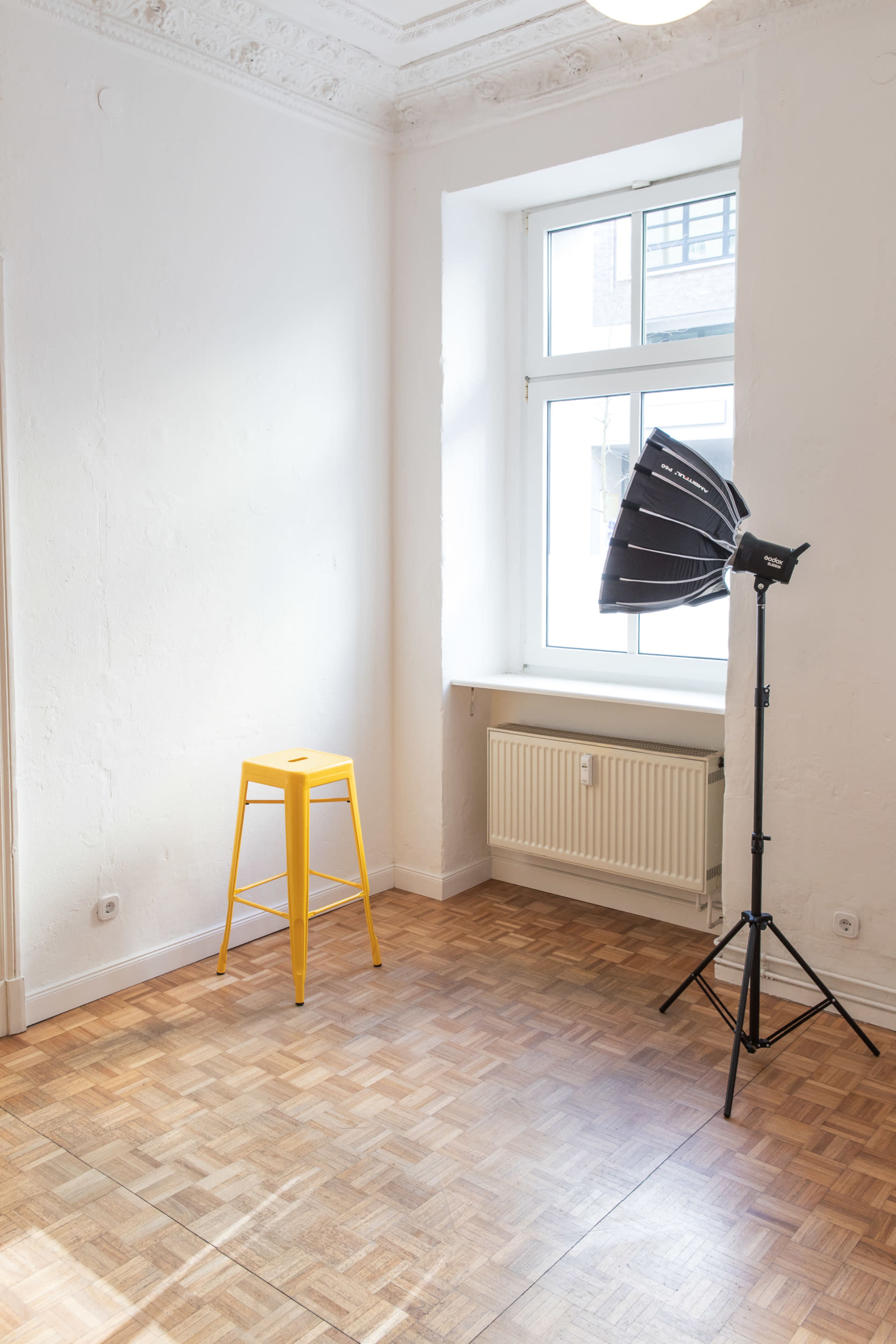 The image shows a bright, empty room with a patterned wooden floor, a yellow bar stool by a window, and a photography light setup in the corner.
