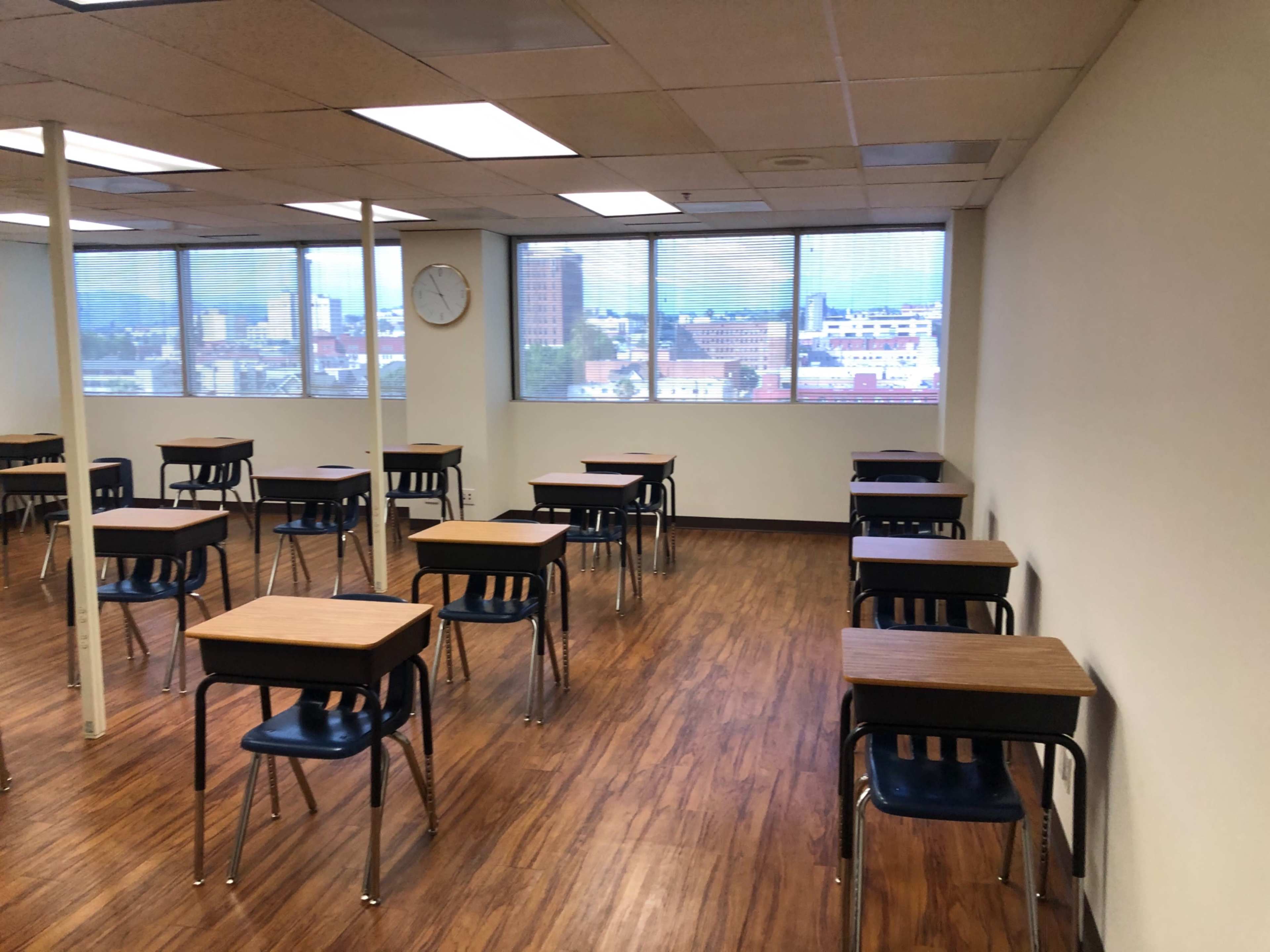 The image shows a classroom with several empty desks arranged in rows and large windows overlooking a cityscape.