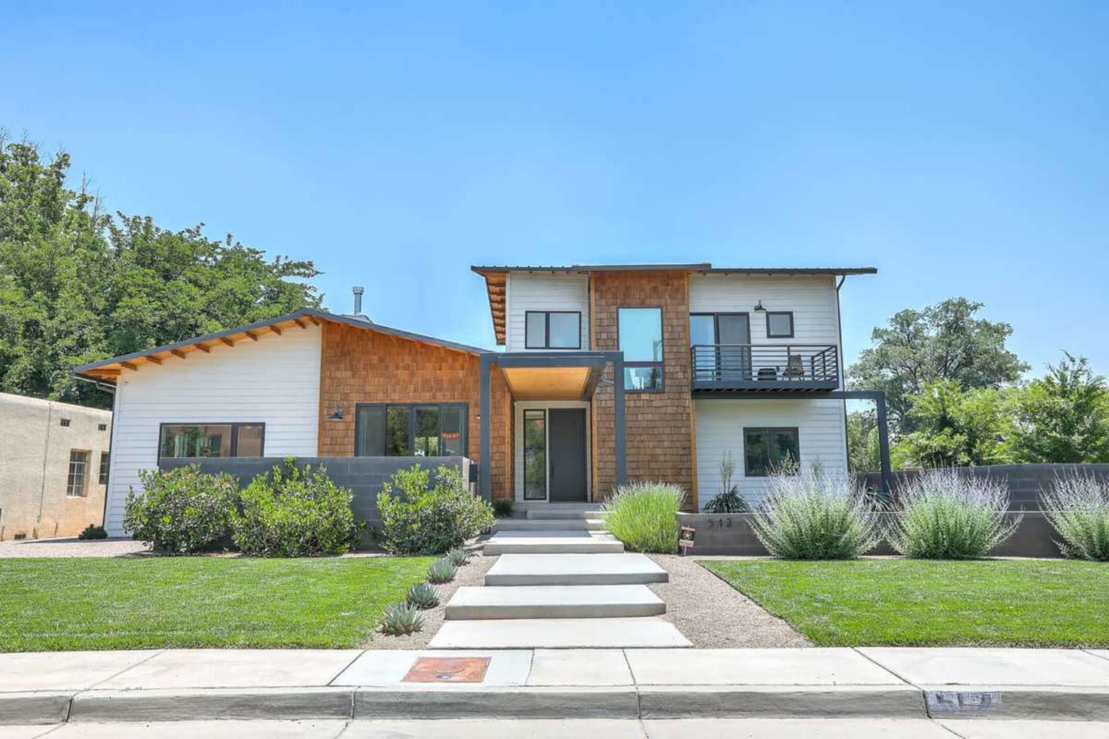 A modern two-story house features a mix of wood and white siding, with landscaped front gardens and a pathway leading to the entrance.
