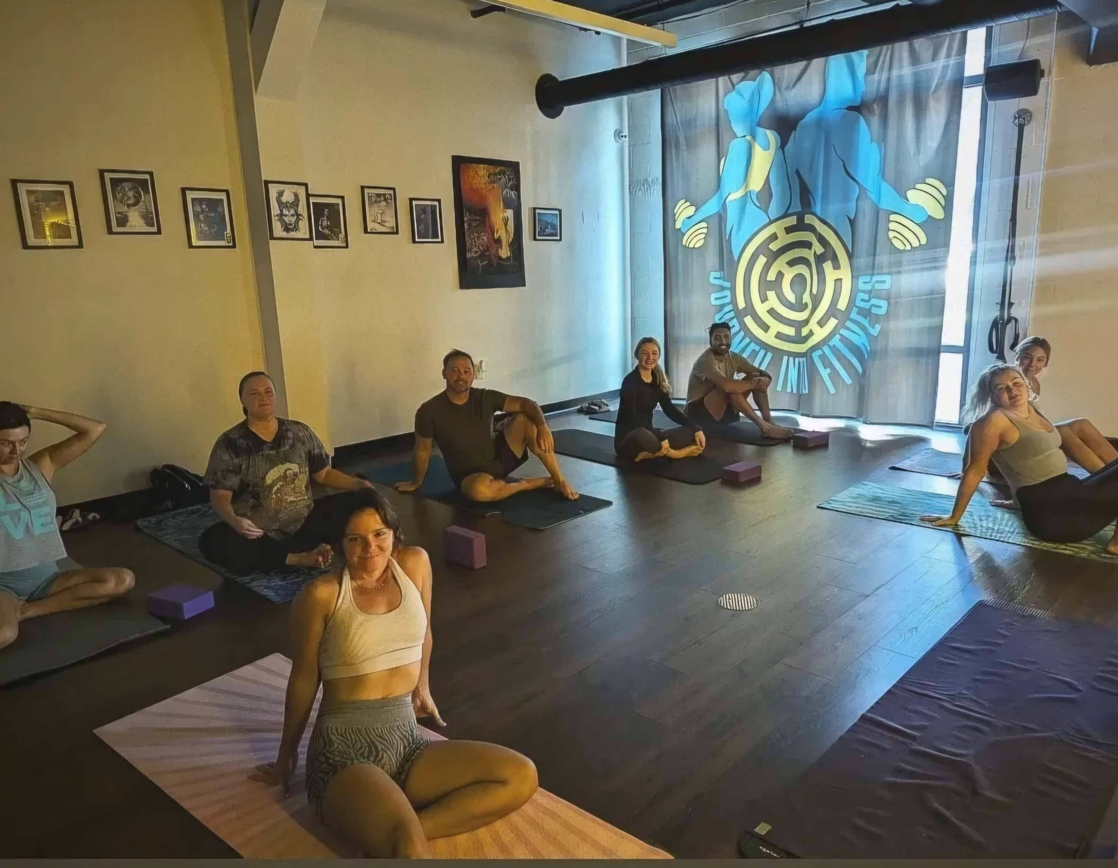 A group of people is seated on yoga mats in a brightly lit studio, preparing for a yoga session, with exercise props placed around them.