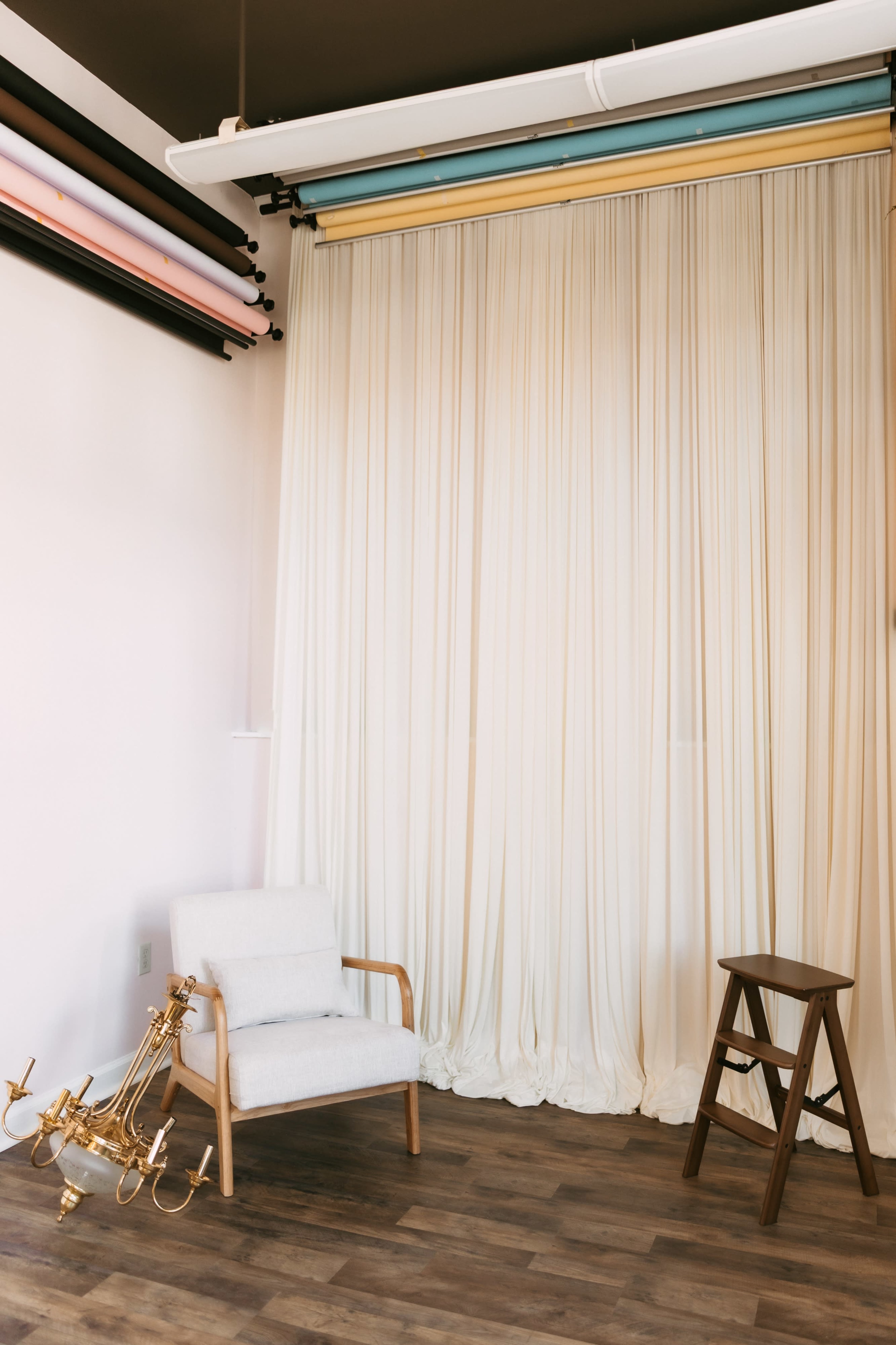 A wooden chair and a stool are positioned near a backdrop of draped fabric in a spacious room with hardwood flooring.