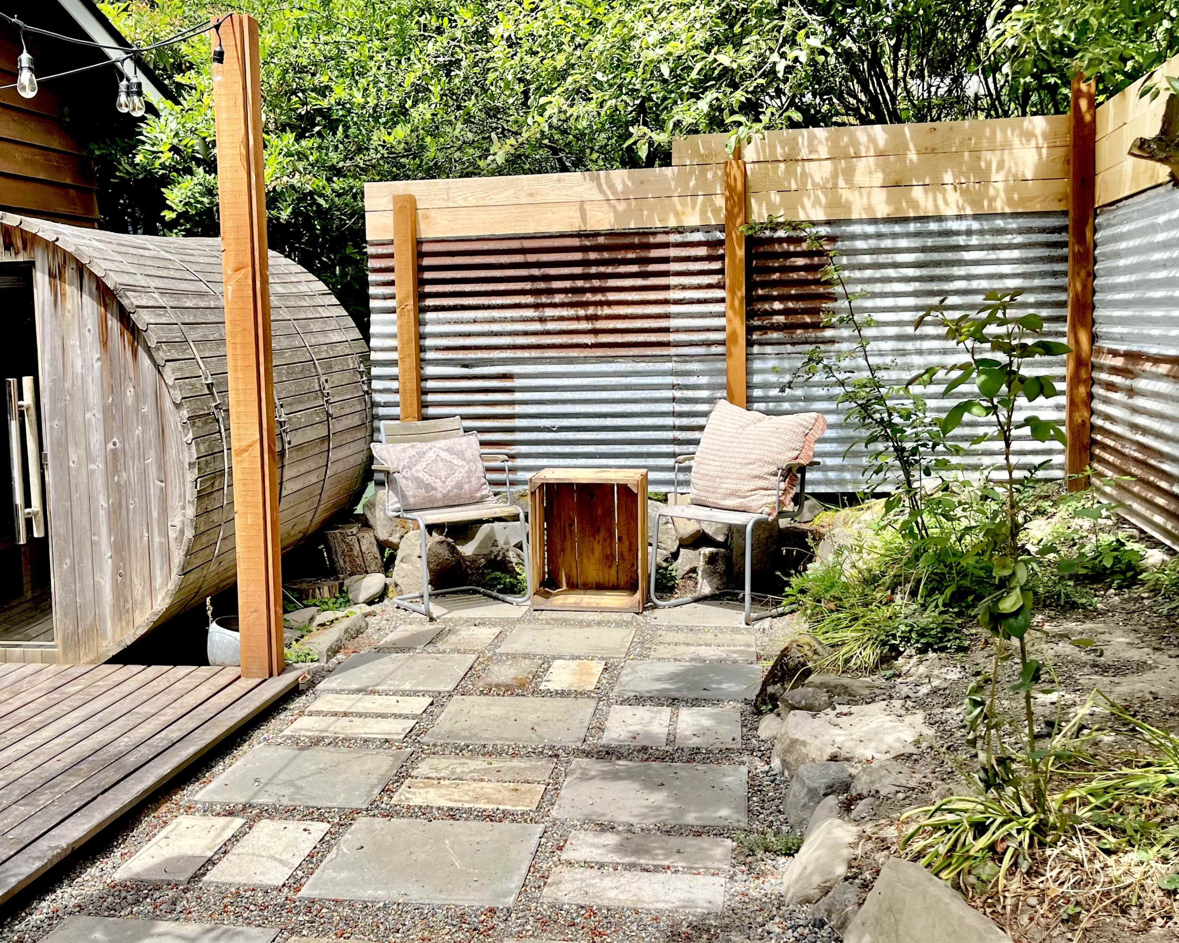 The image shows a small outdoor patio area with a circular wooden structure, two chairs, and a wooden table surrounded by stone and gravel paths, with plants and corrugated metal fencing.