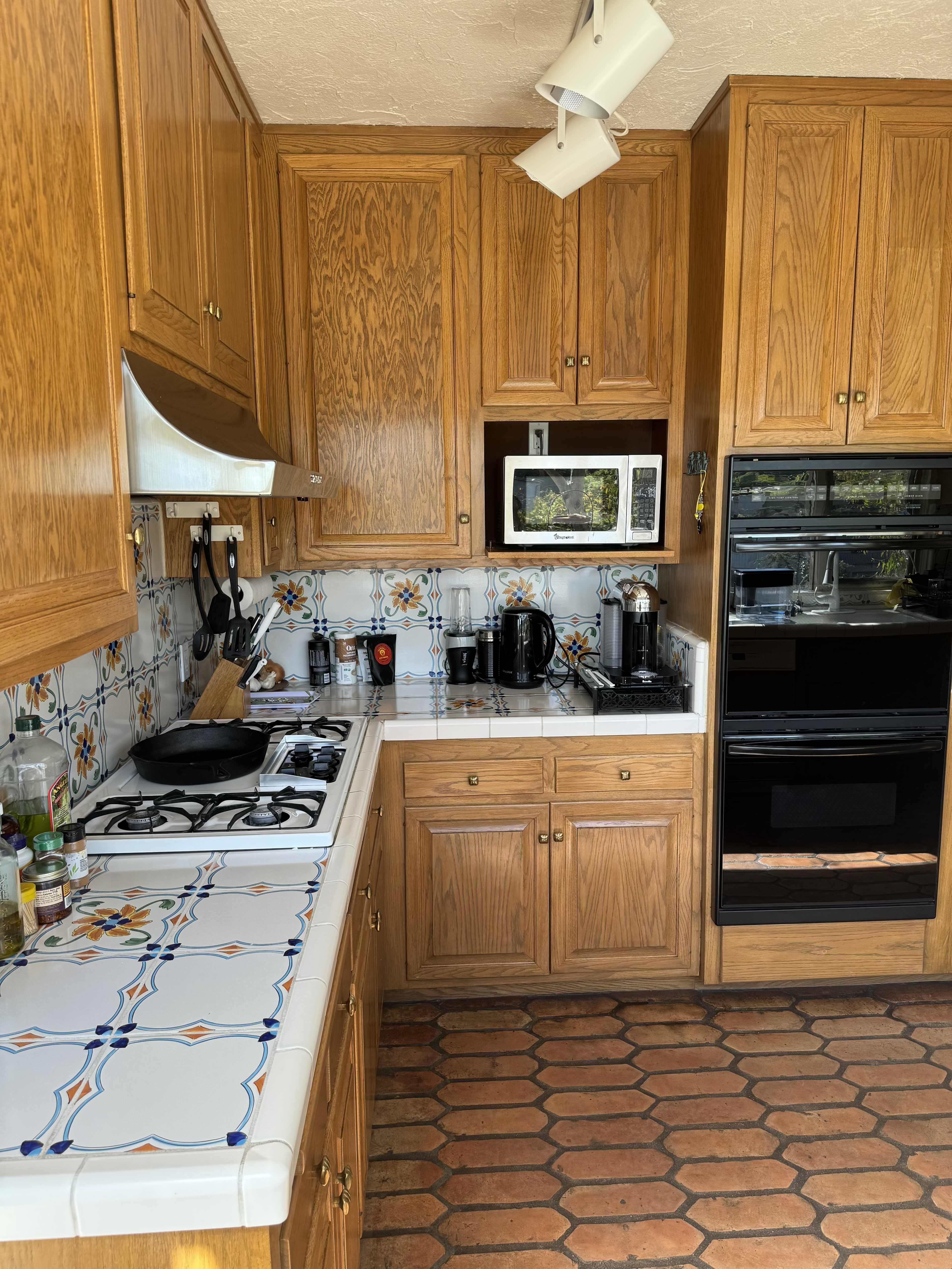 A kitchen with wooden cabinets, tiled countertops, a gas stove, a microwave, and an oven, featuring a patterned backsplash and a tiled floor.