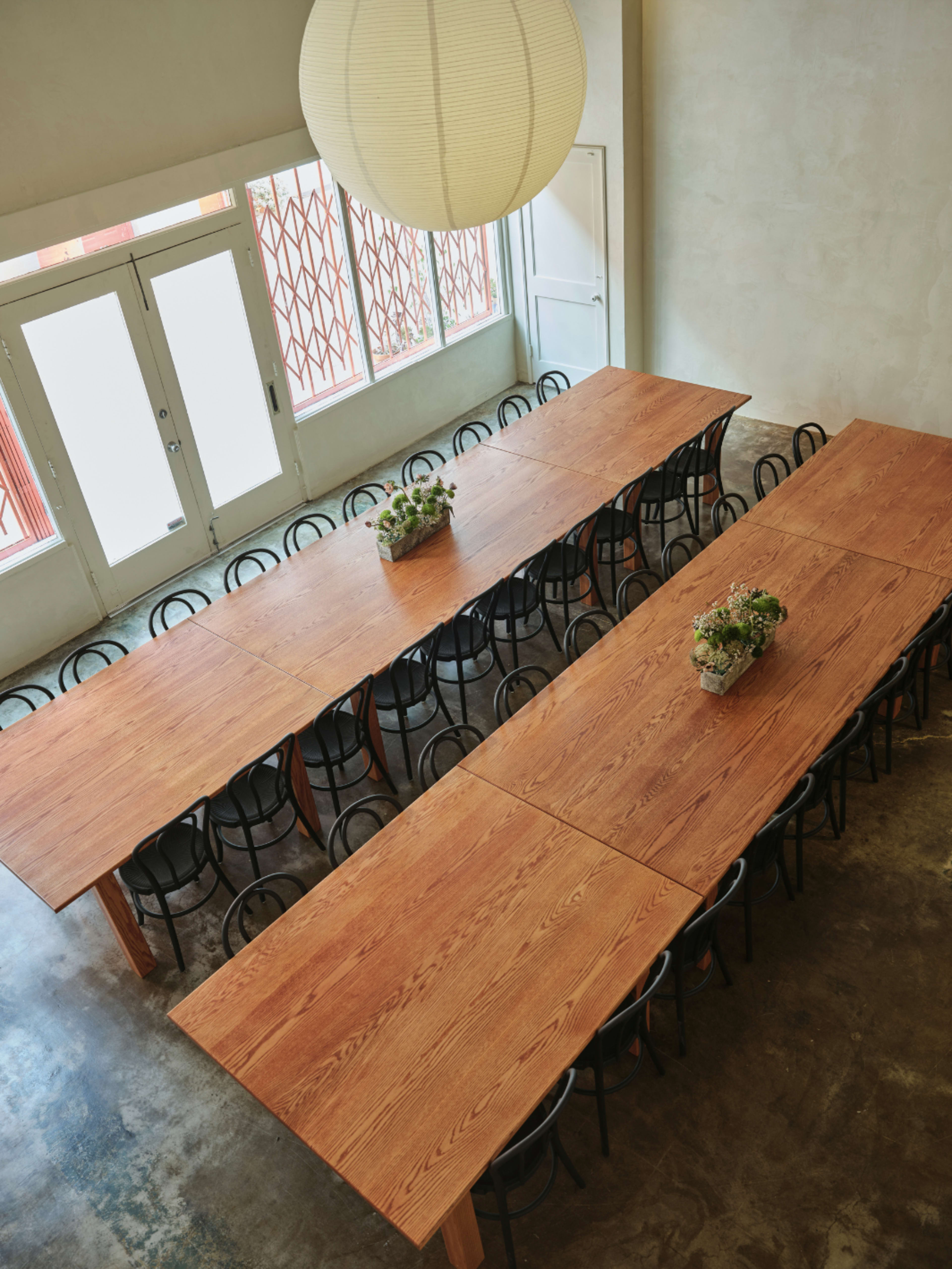 The image shows a spacious room with two long wooden tables arranged in parallel, surrounded by black chairs, under a large paper lantern and with large windows allowing natural light to enter.