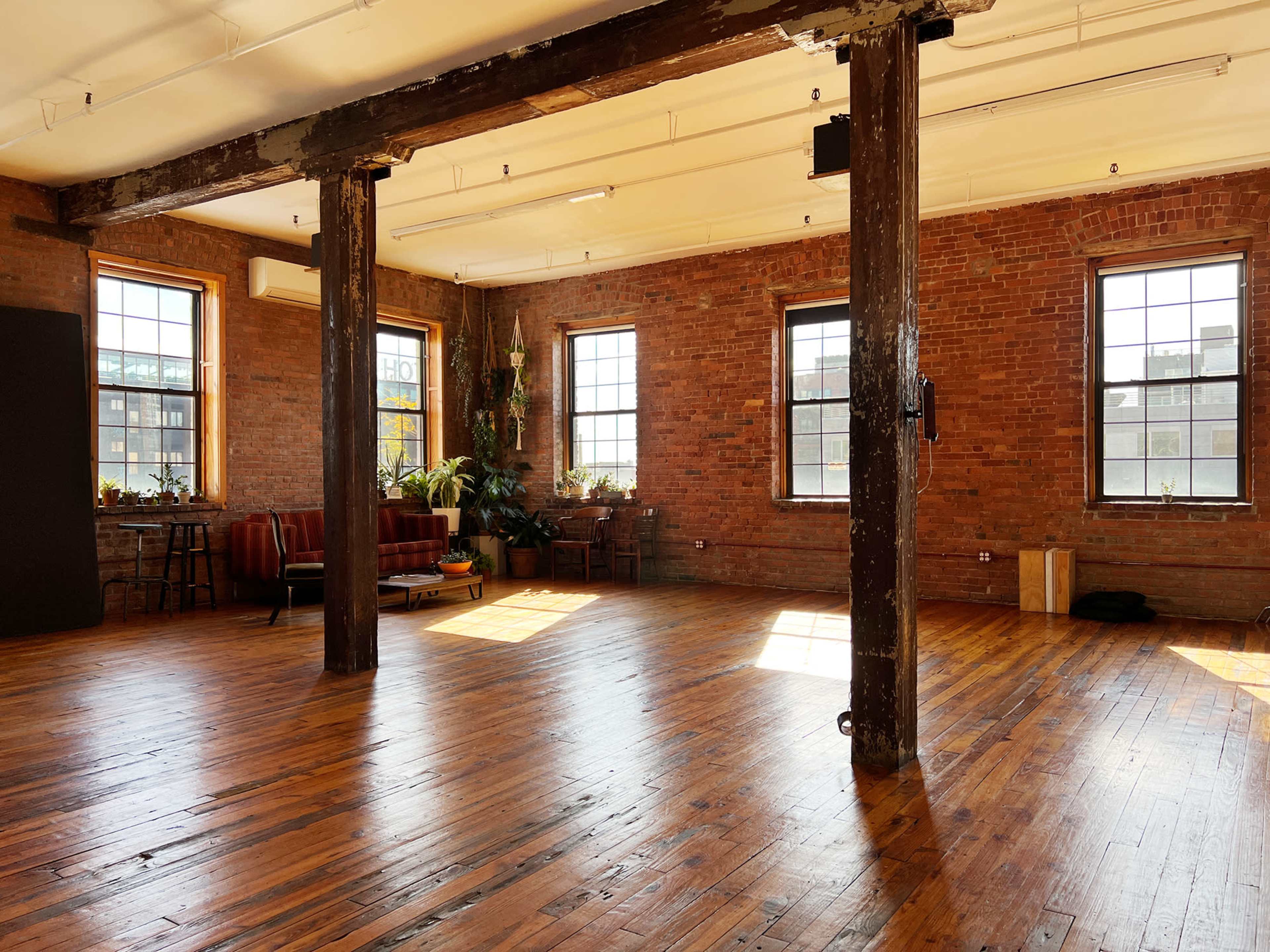A spacious interior with wooden floors, large windows, brick walls, and a seating area with a sofa and plants.