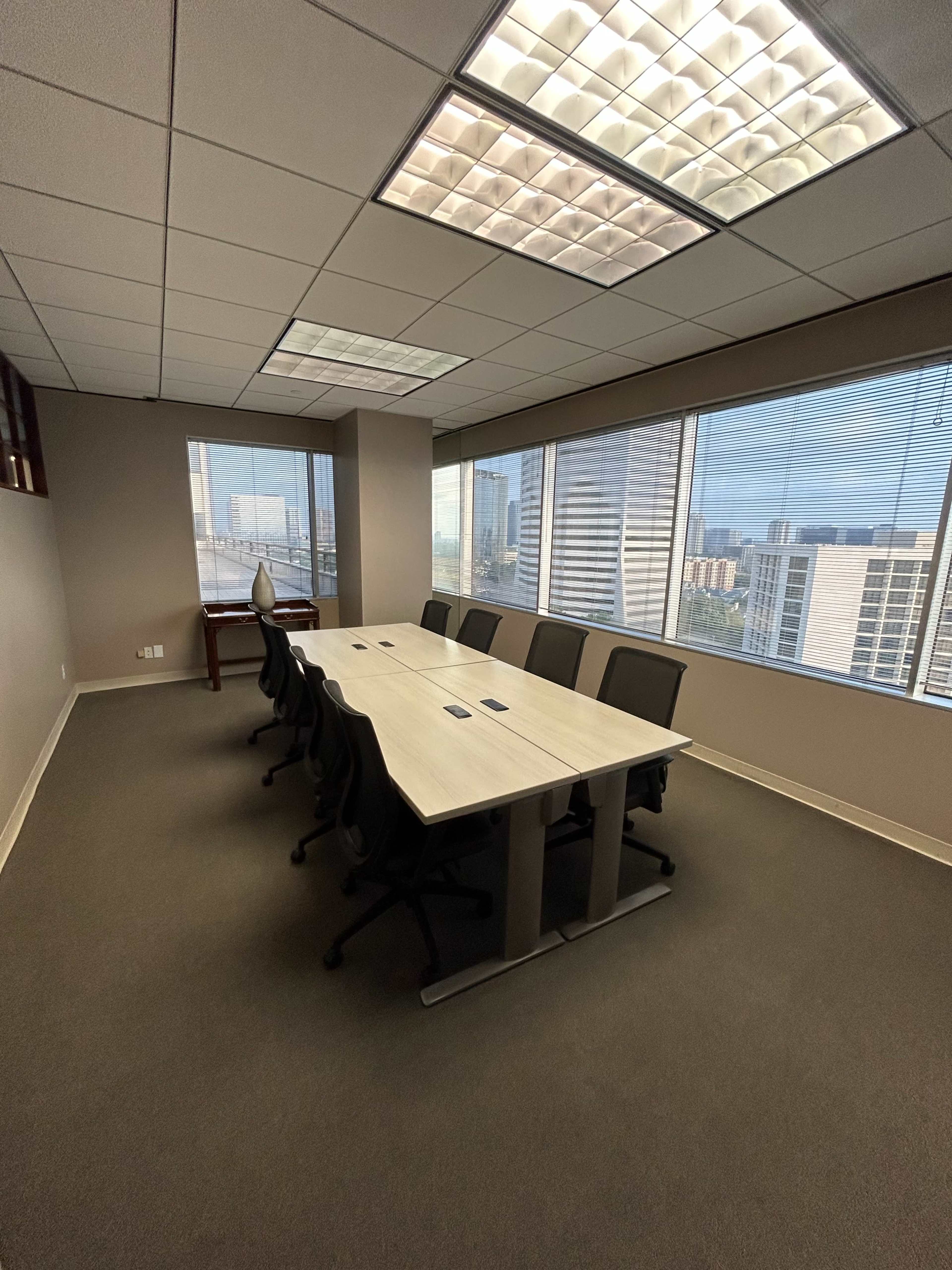 A modern conference room features a long table, seven ergonomic chairs, and large windows with a view of the city skyline.