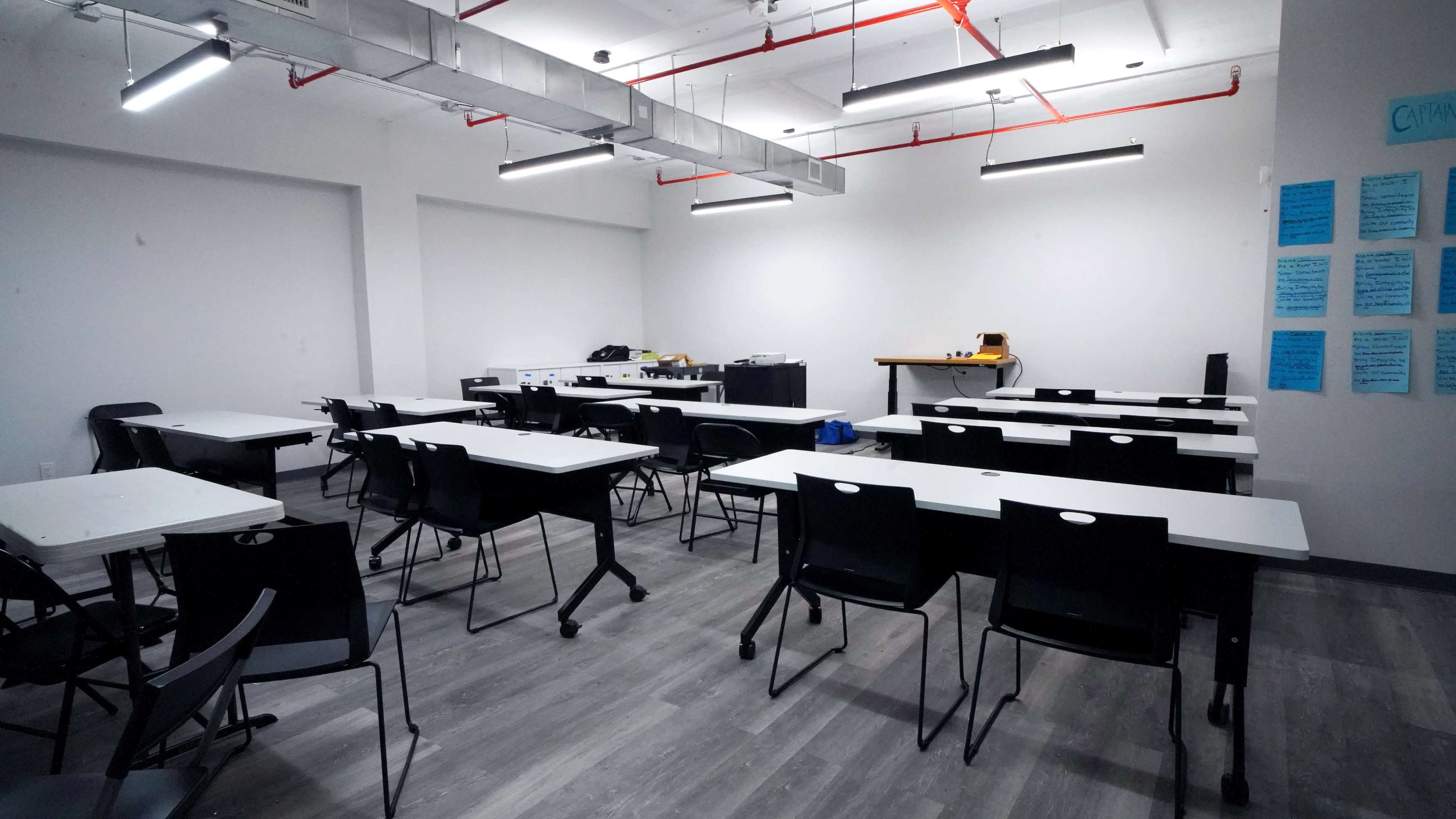 A nearly empty classroom with rows of black chairs and white tables, illuminated by overhead lights.