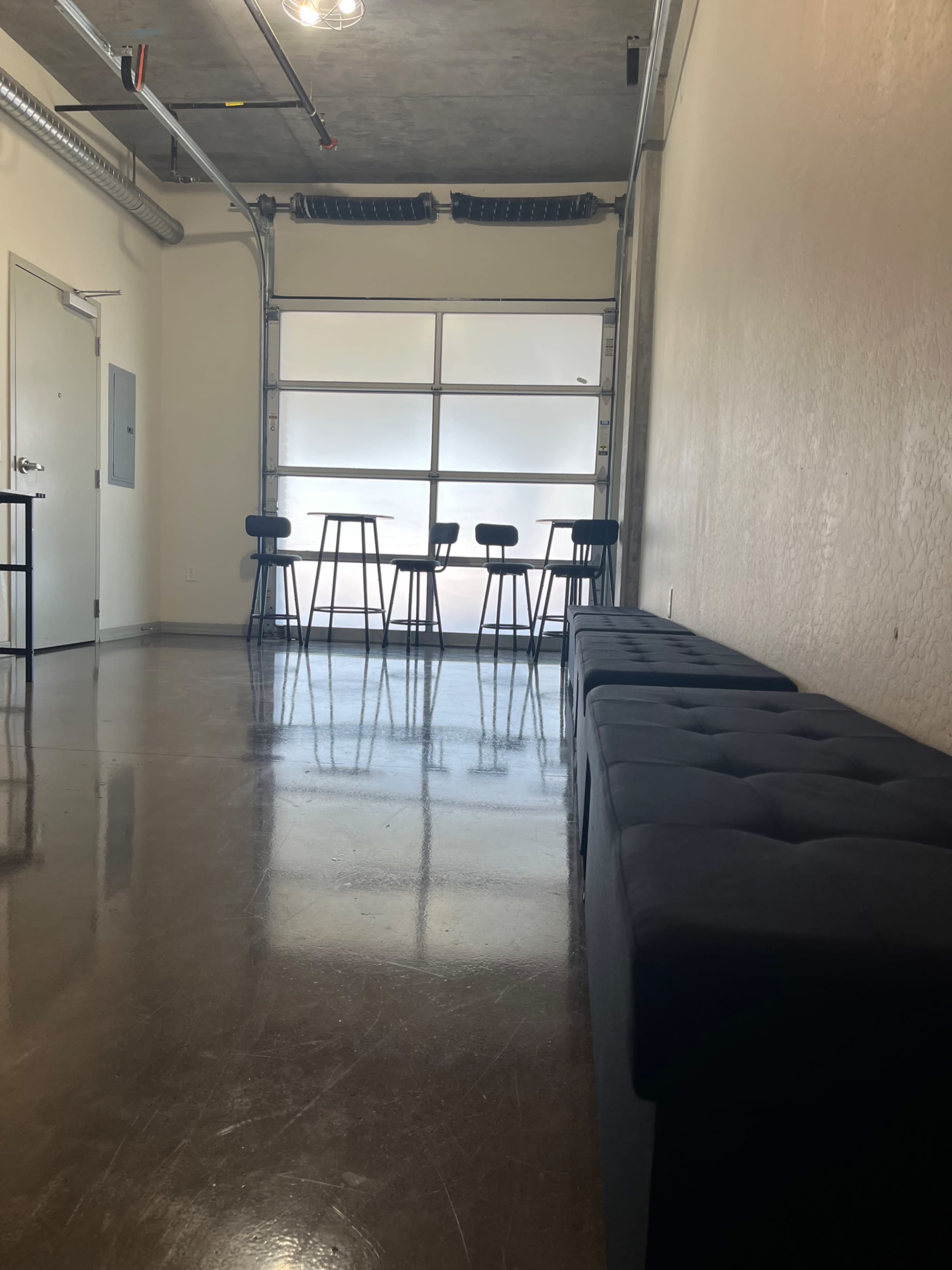 The image shows a narrow room with polished concrete flooring, featuring a line of black benches on one side and a row of tall stools with a window in the background.
