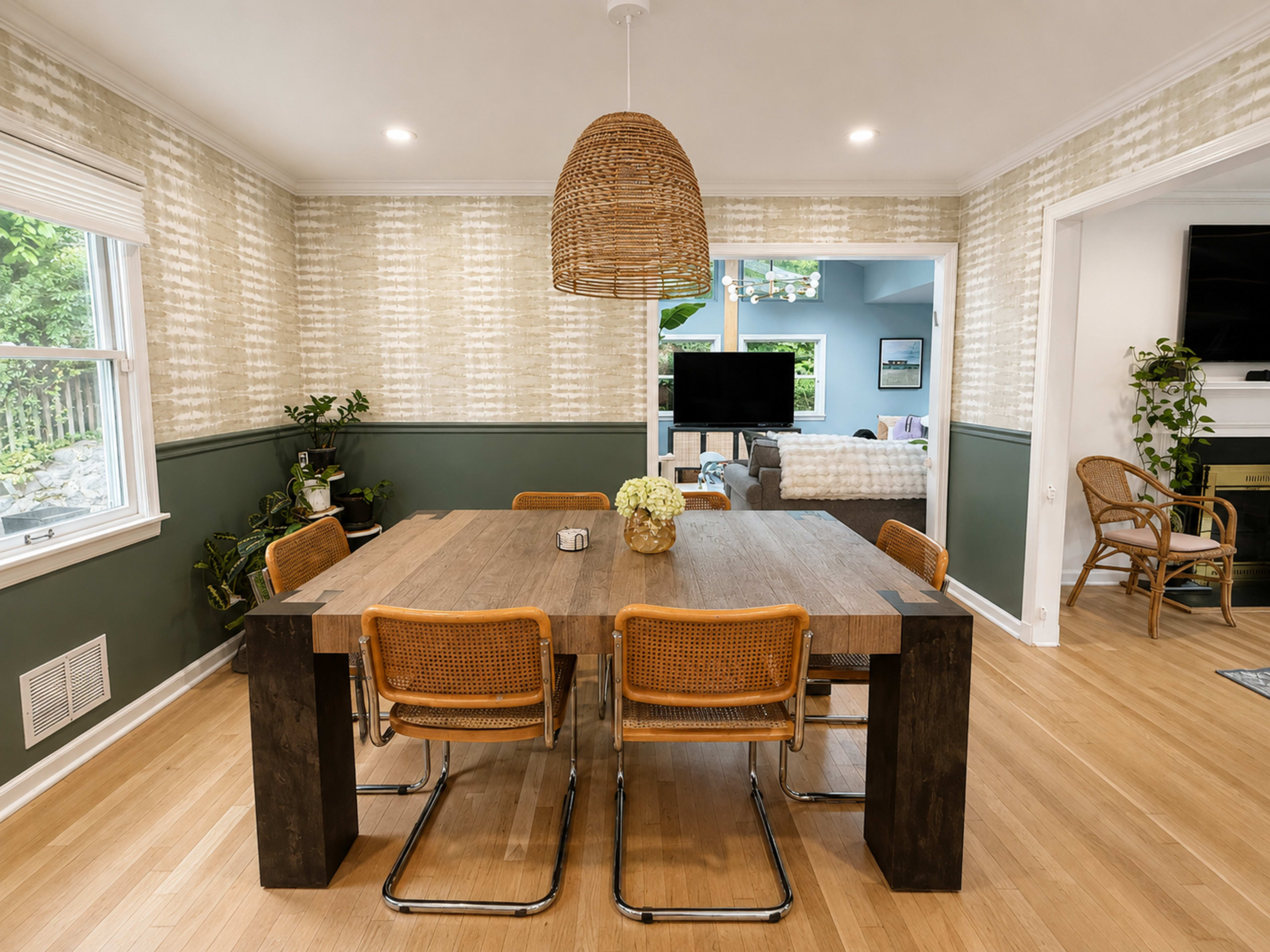 The image shows a dining room with a large wooden table surrounded by six rattan chairs, accented by a woven pendant light and green-patterned walls.