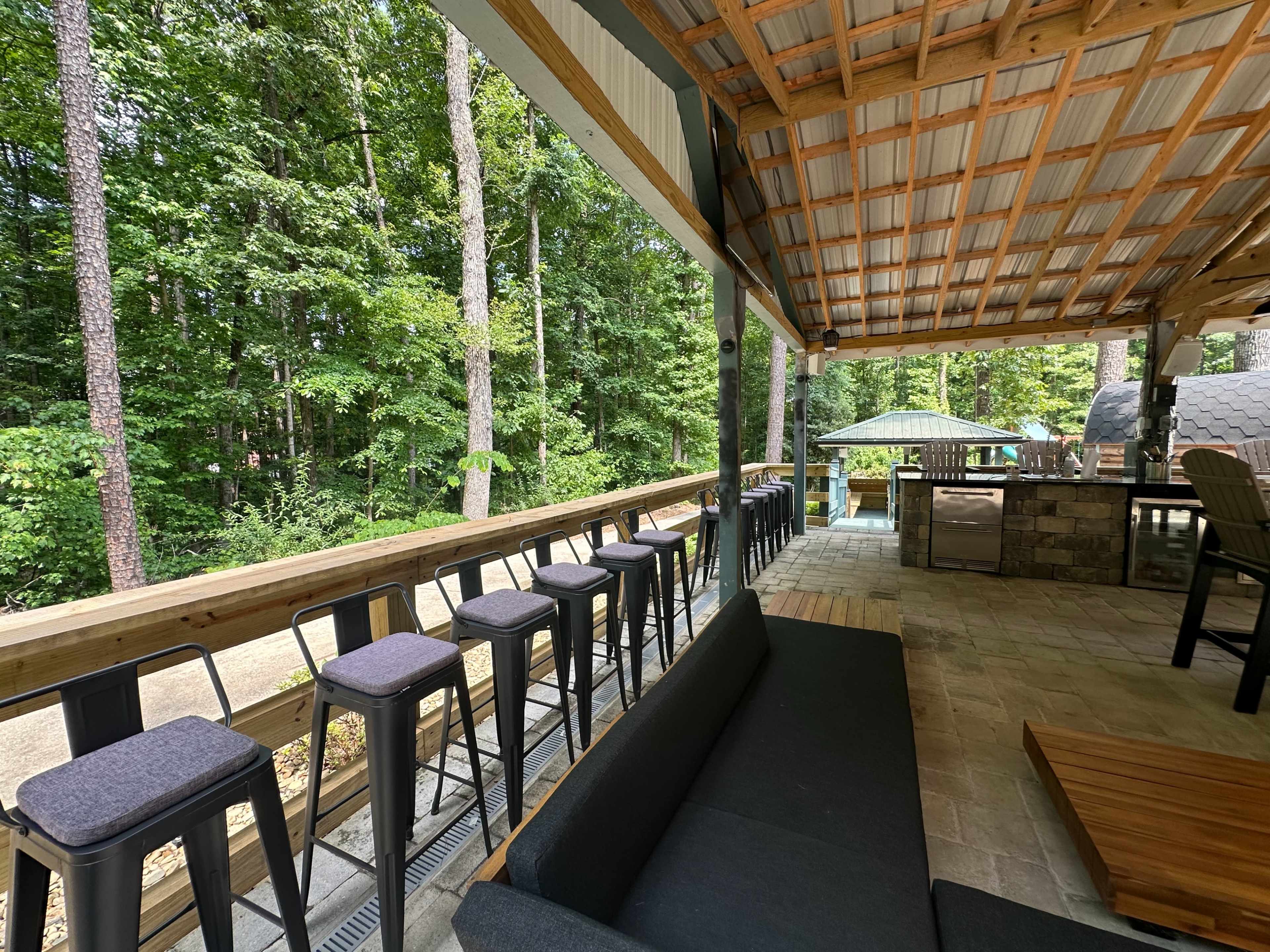 A covered outdoor patio area with a row of tall stools facing a natural wooded backdrop.