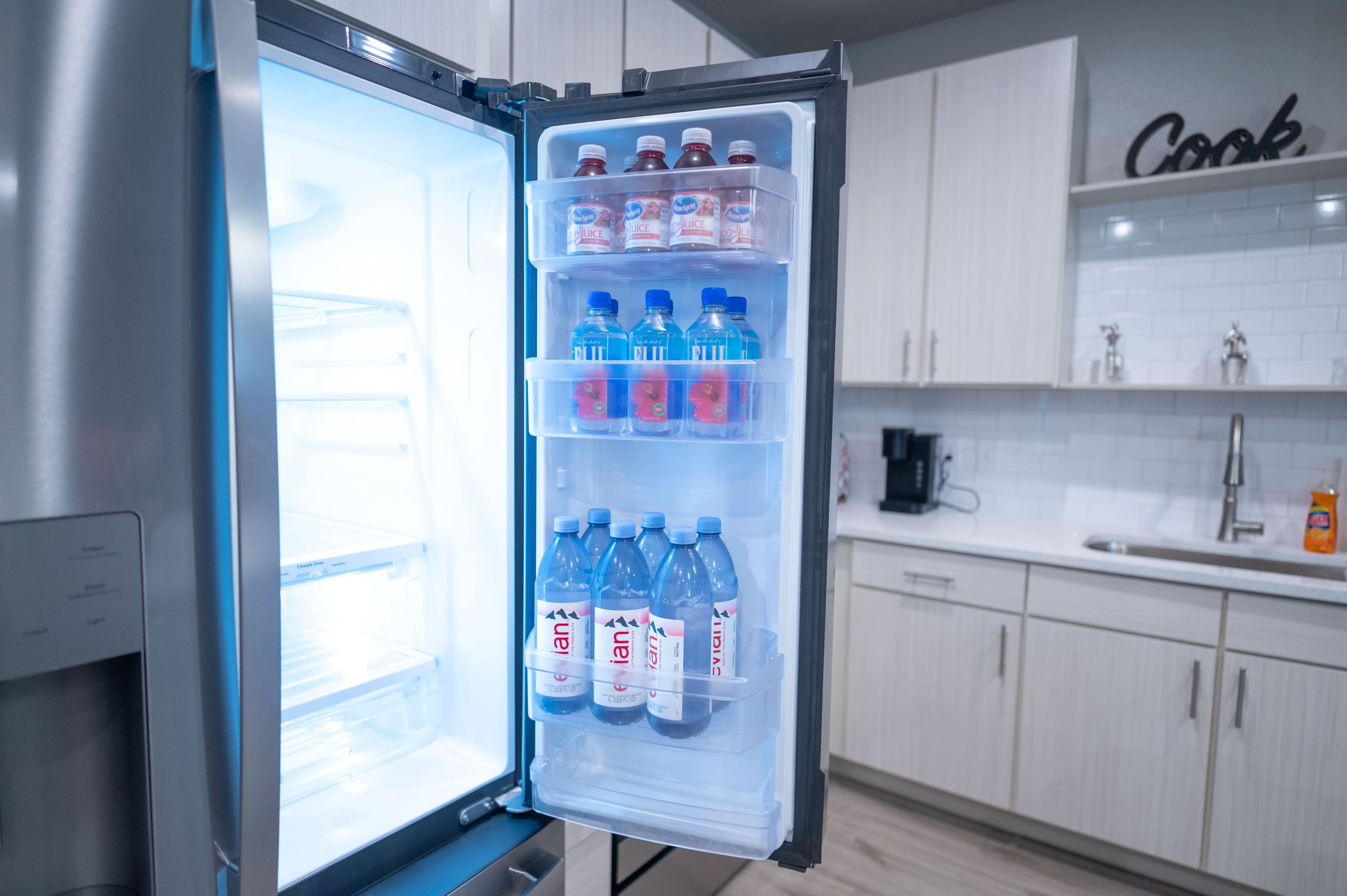 The image shows the interior of a refrigerator with shelves containing bottles of water and flavored drinks, alongside a bright kitchen backdrop.