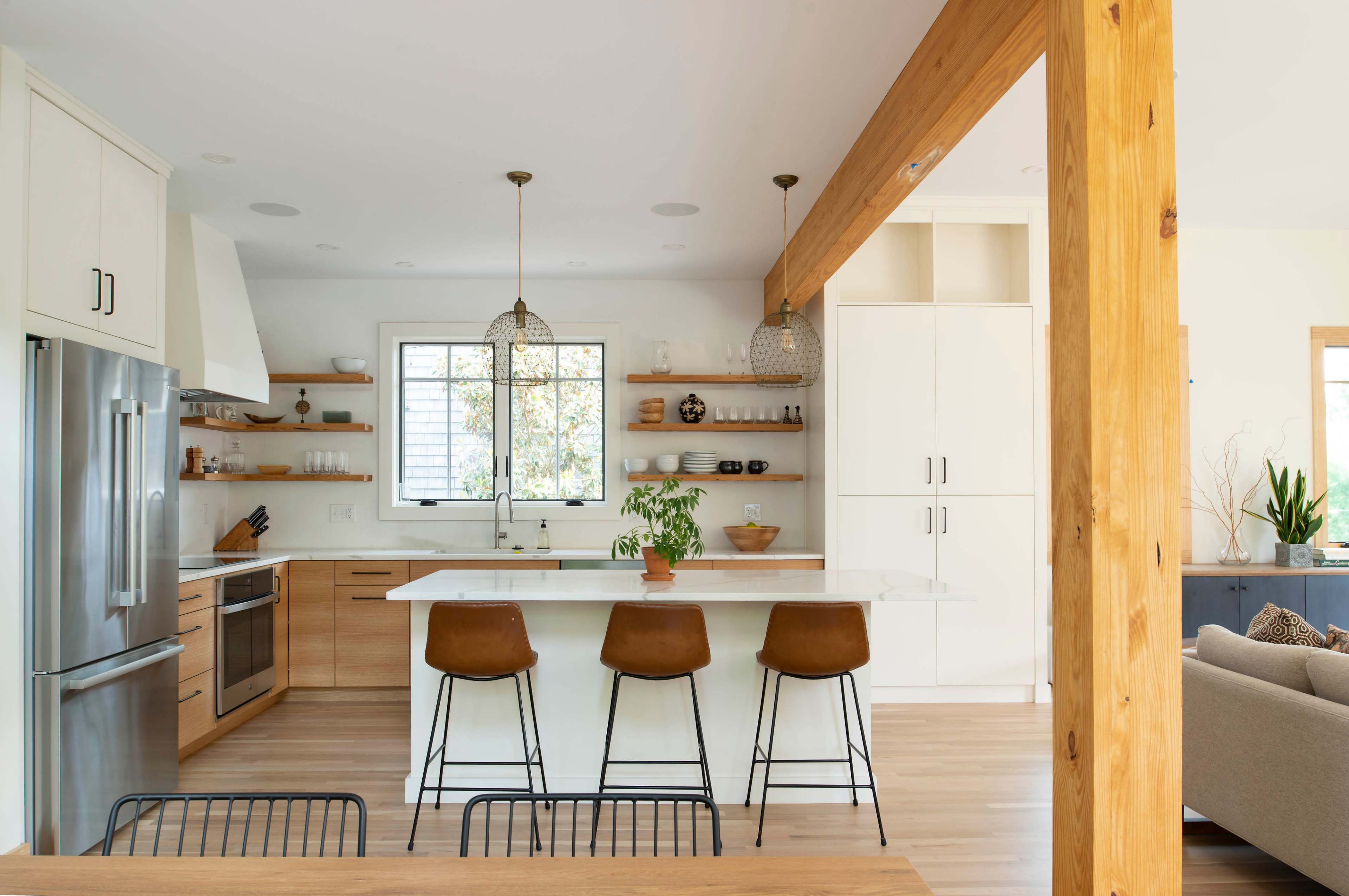 A modern kitchen features a central island with three brown bar stools, wooden shelves, and white cabinetry, illuminated by pendant lights.