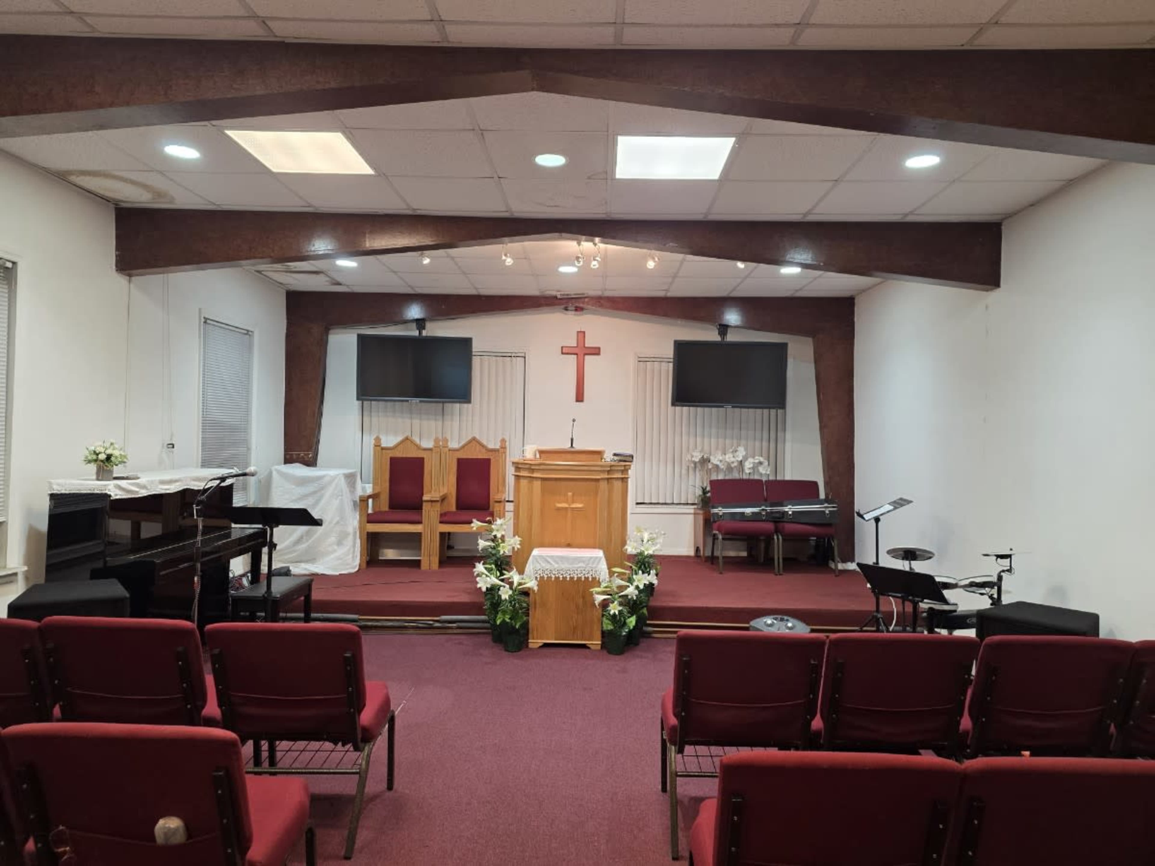 The interior of a church features a wooden podium at the front, flanked by two large screens, with rows of red chairs arranged for seating.