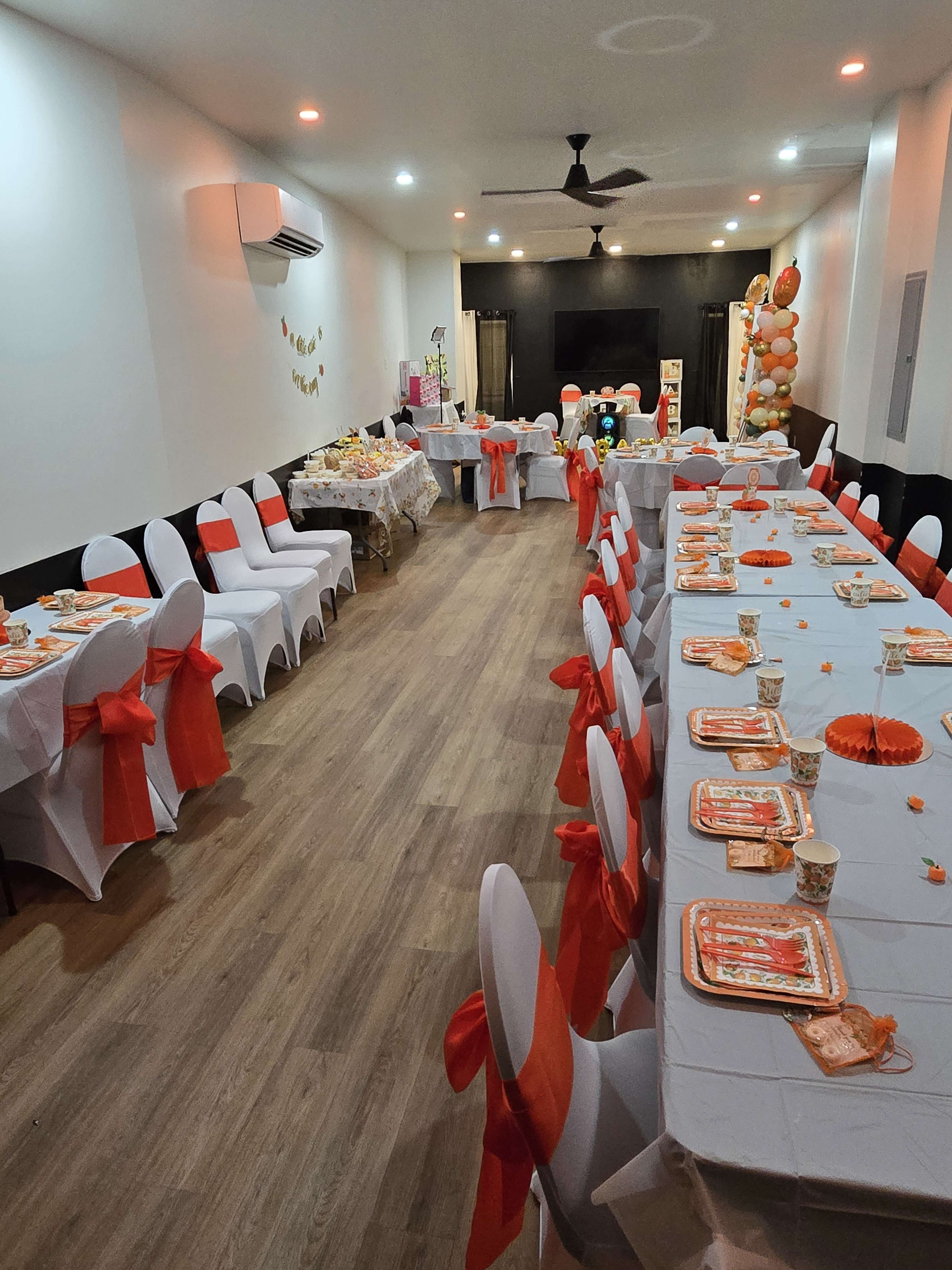 The image shows a banquet hall set up for a party, with tables covered in white cloths and orange sashes, arranged in rows and decorated with festive tableware.