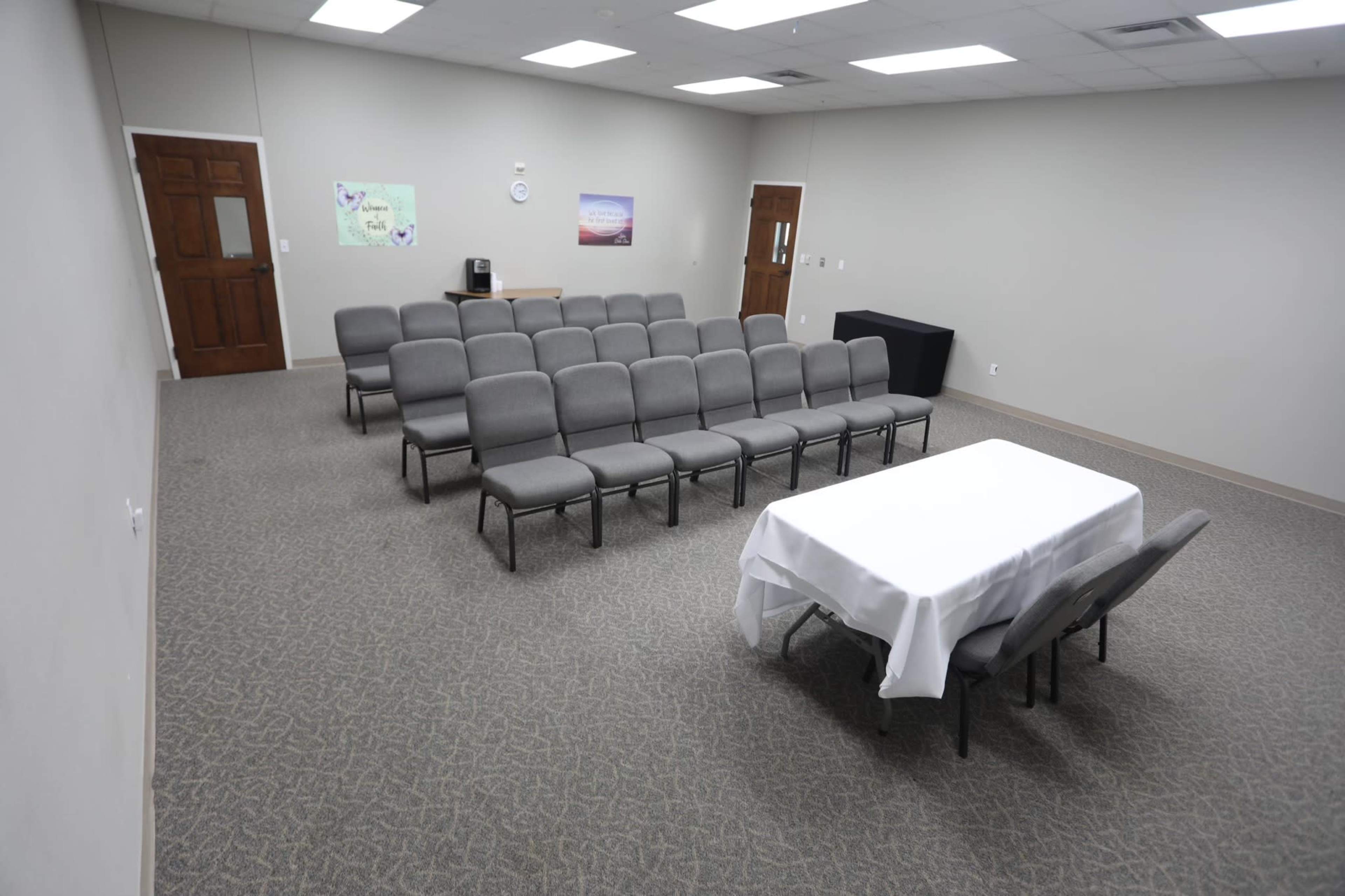 The image shows a sparsely furnished conference room with rows of gray chairs and a table covered with a white tablecloth at the front.