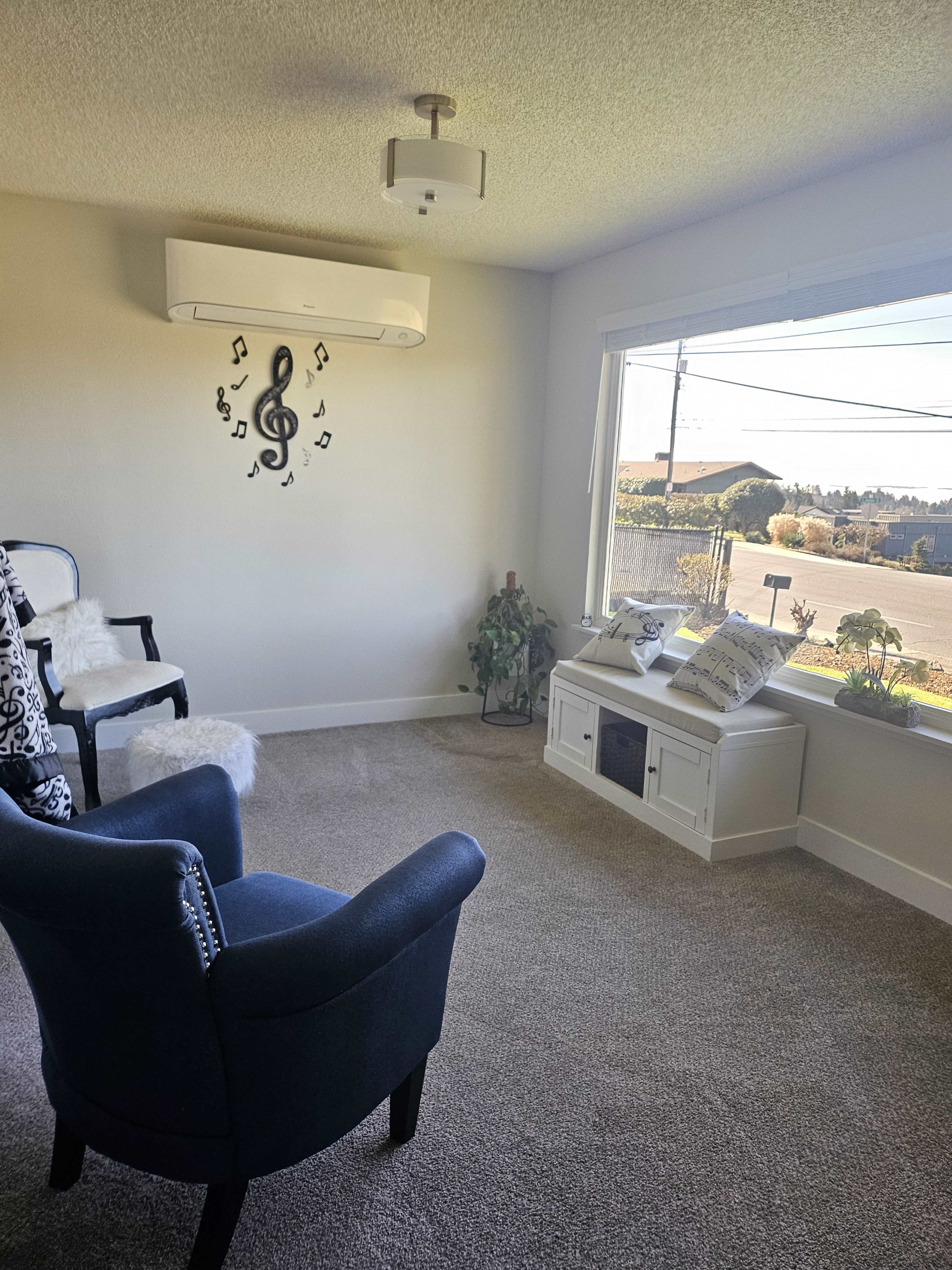 The image shows a bright living room with a large window, two chairs, a music-themed wall decoration, and a small cabinet beneath the window.