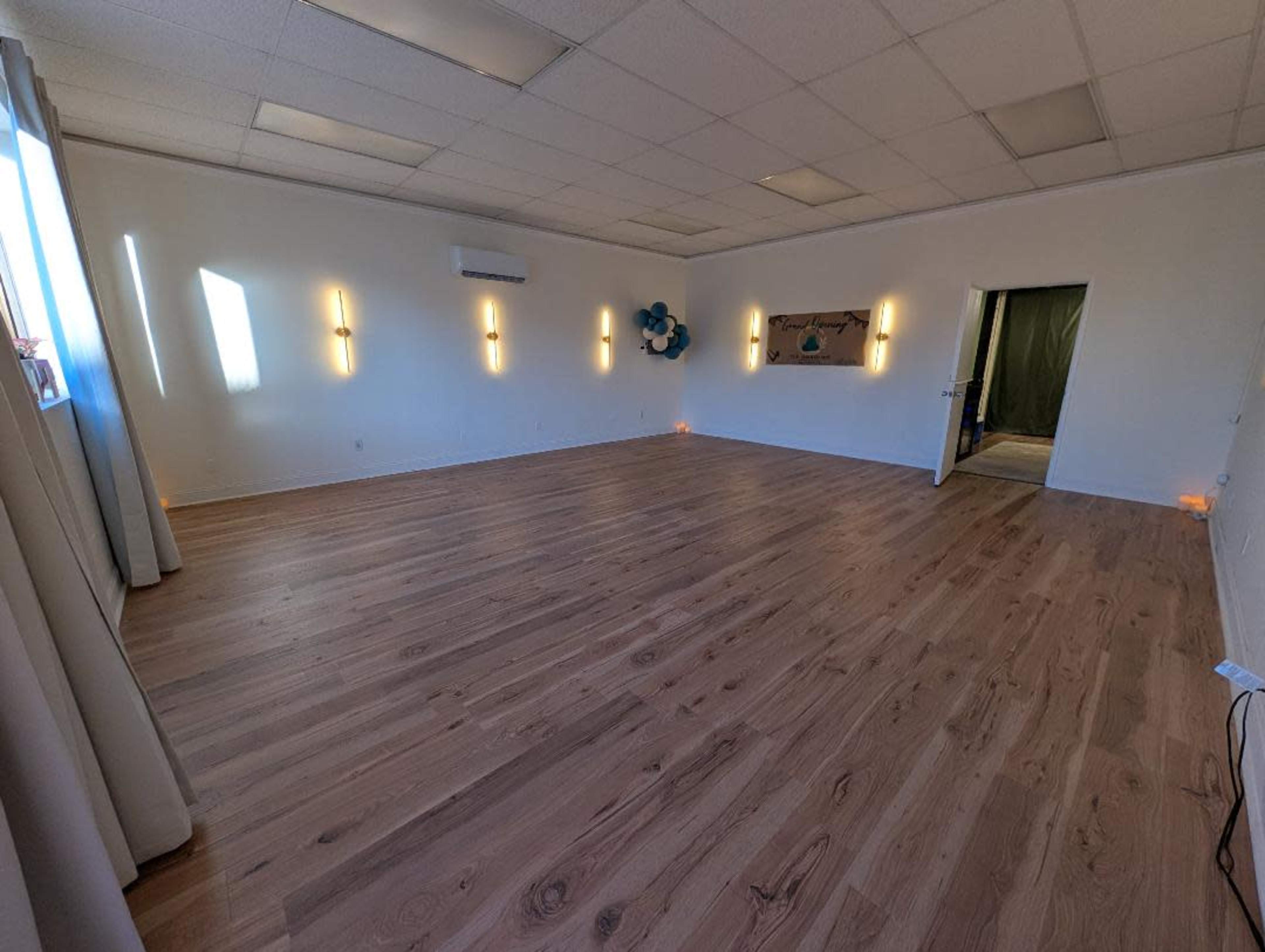 The image shows a spacious, empty room with wooden flooring, well-lit by wall sconces and large windows.