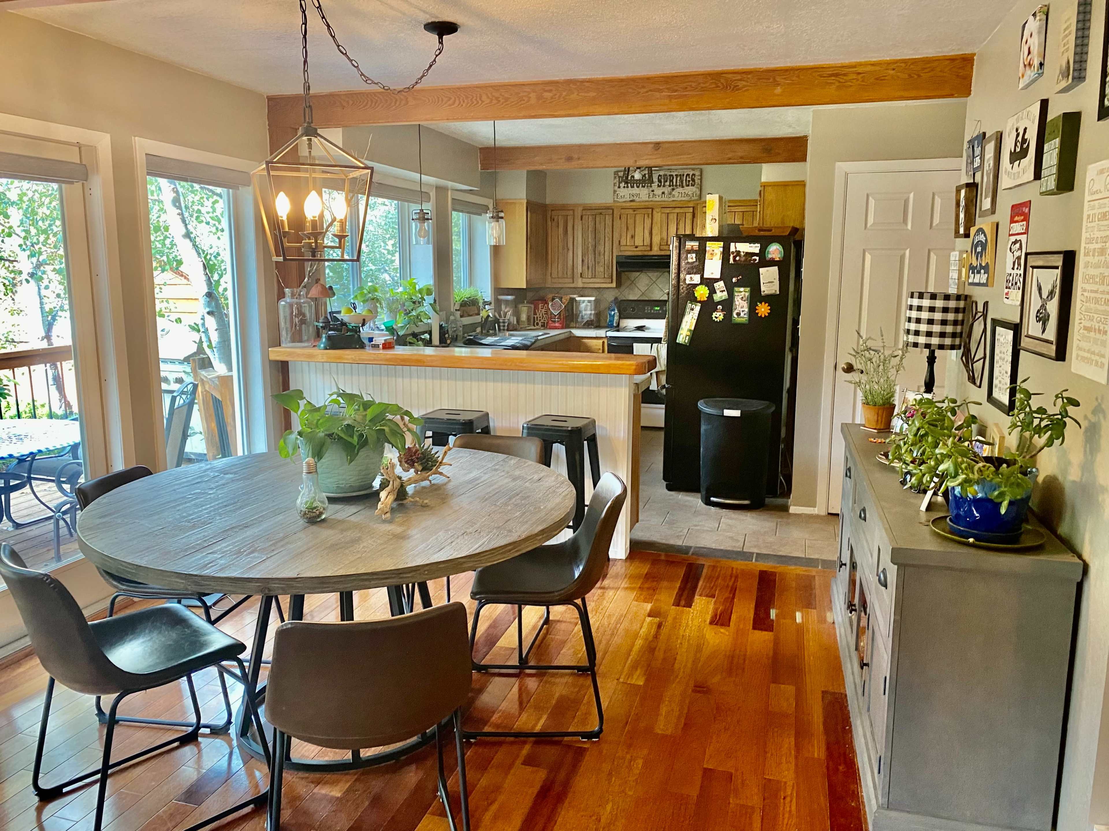 A round dining table surrounded by chairs is positioned in a bright kitchen area with wooden floors and a view of the outdoors through large windows.