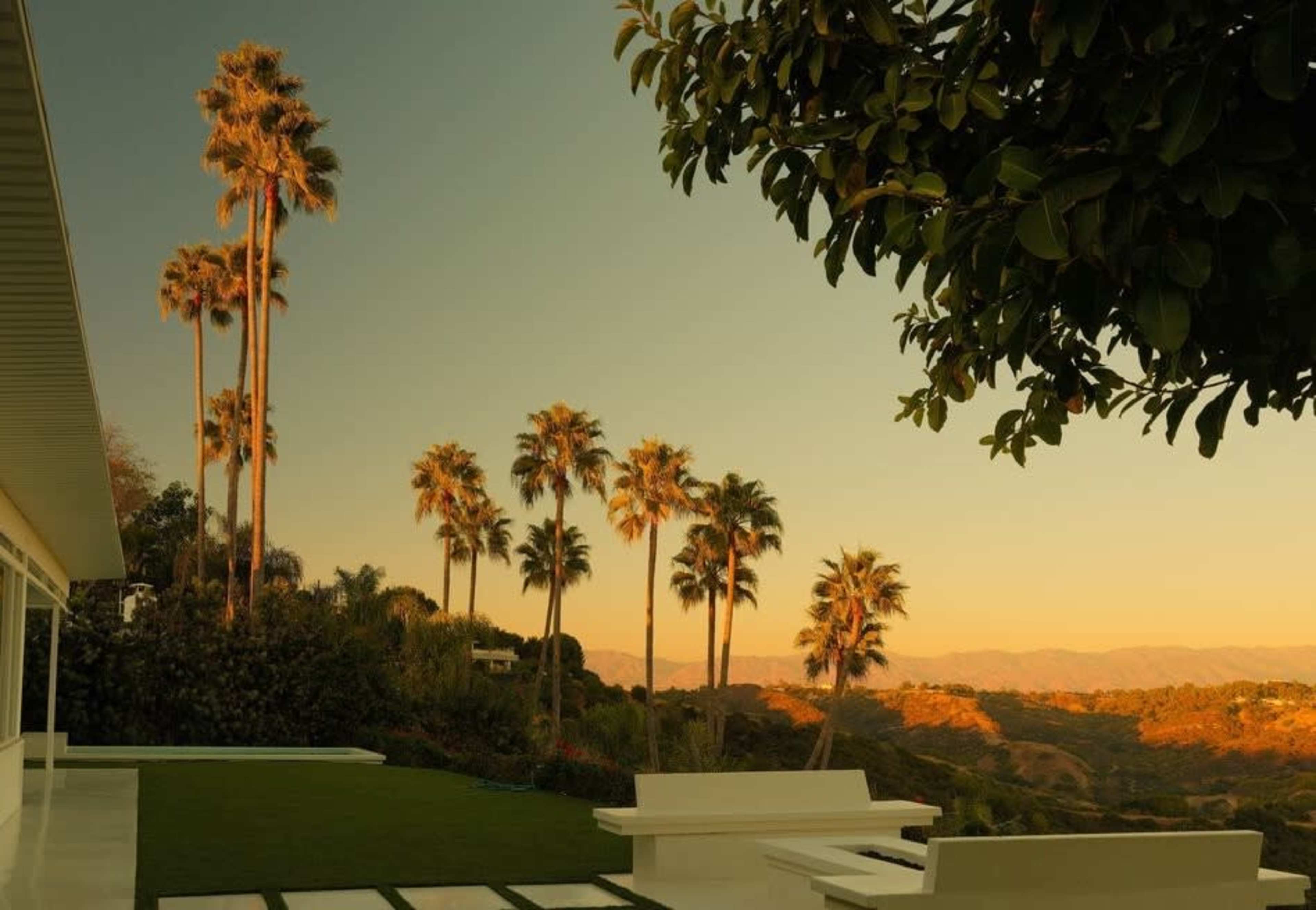 The image shows a modern outdoor living space with white furniture and several tall palm trees against a backdrop of rolling hills during sunset.
