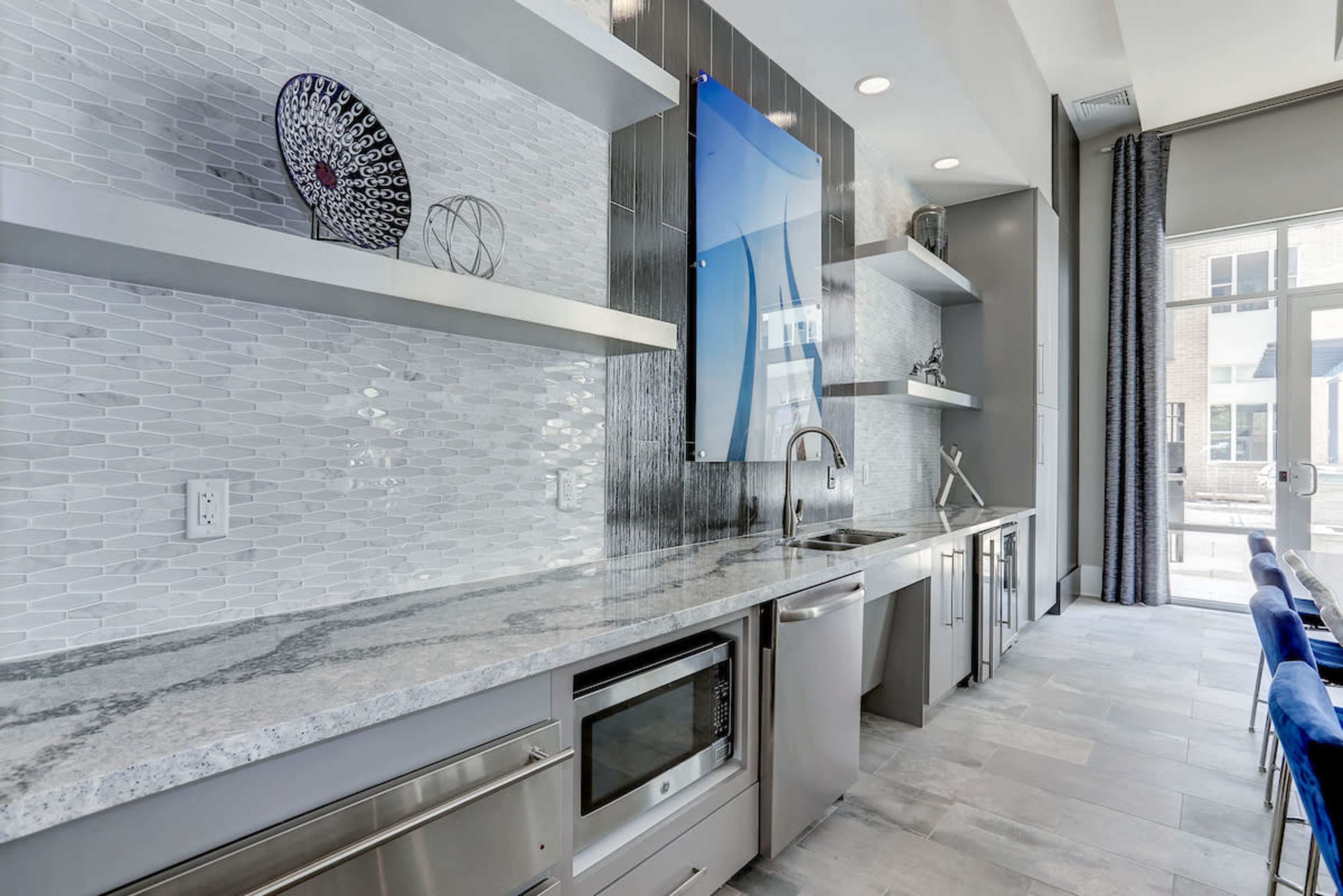 The image shows a modern kitchen with a sleek granite countertop, stainless steel appliances, and a minimalist design featuring open shelves and a decorative wall piece.