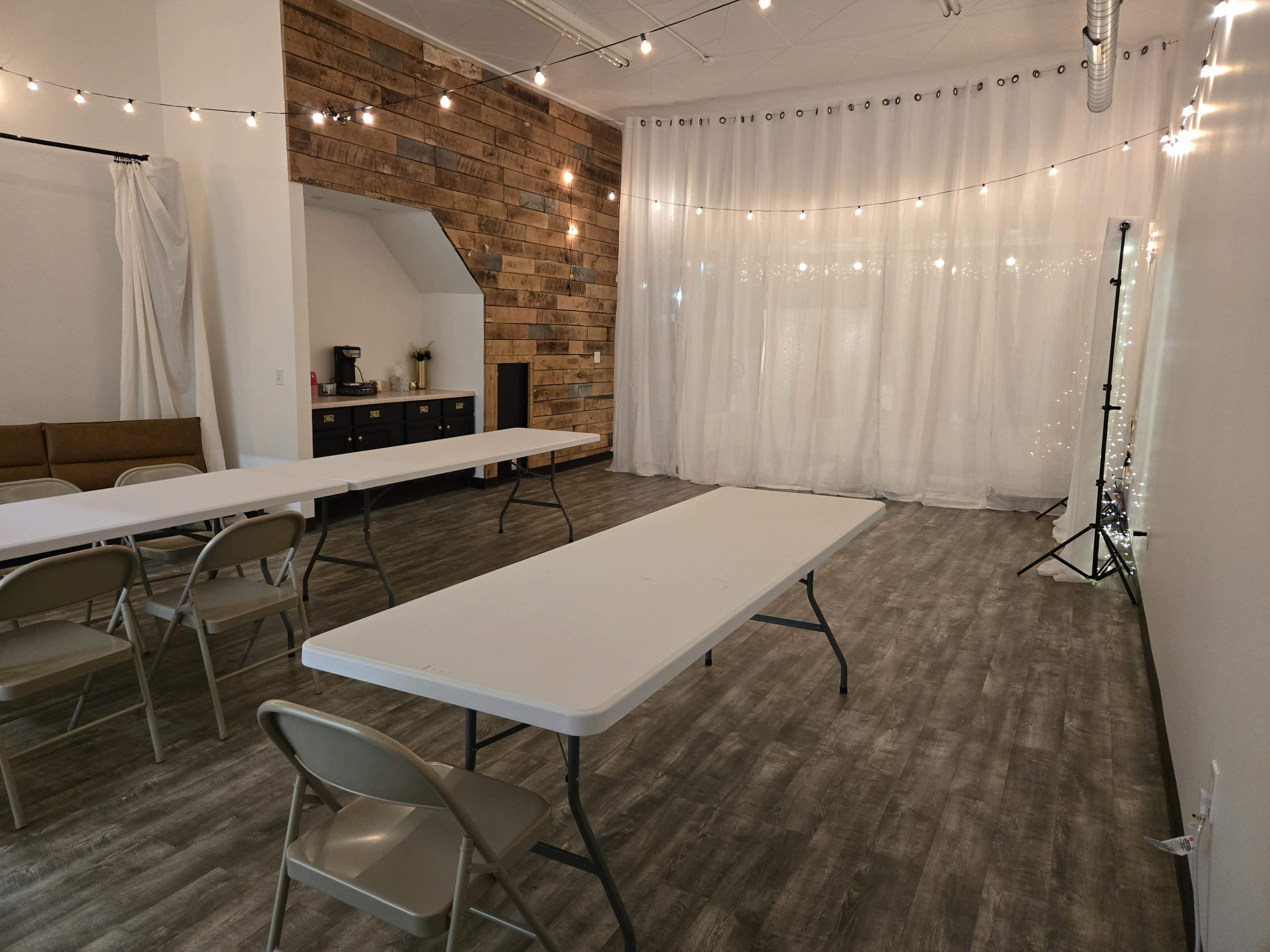 The image shows a spacious room arranged with multiple white tables and folding chairs, featuring a backdrop of string lights and sheer curtains.