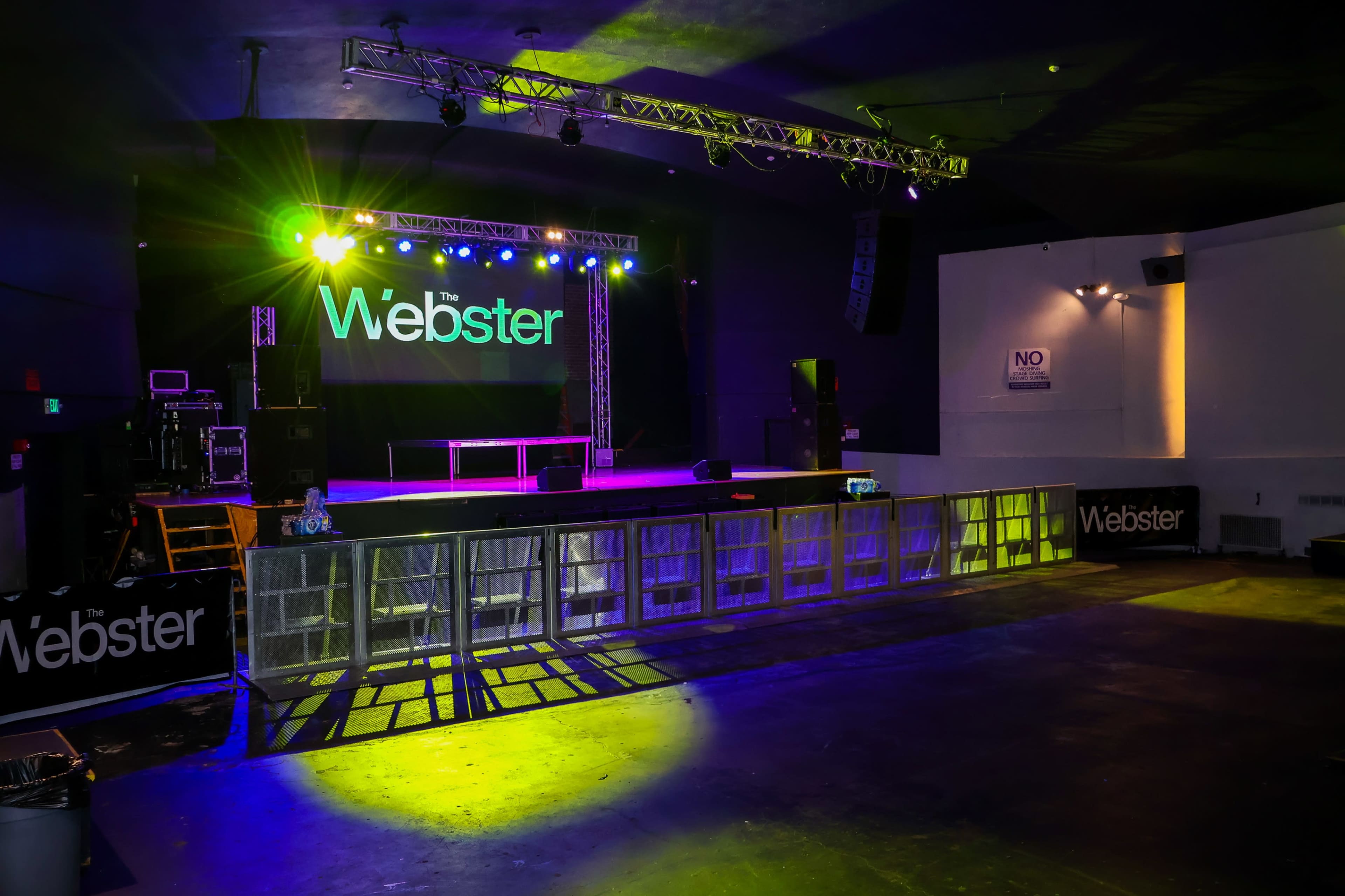 A small concert venue with a stage illuminated by colorful lights, featuring barriers in front of the stage and signage displaying "the Webster."