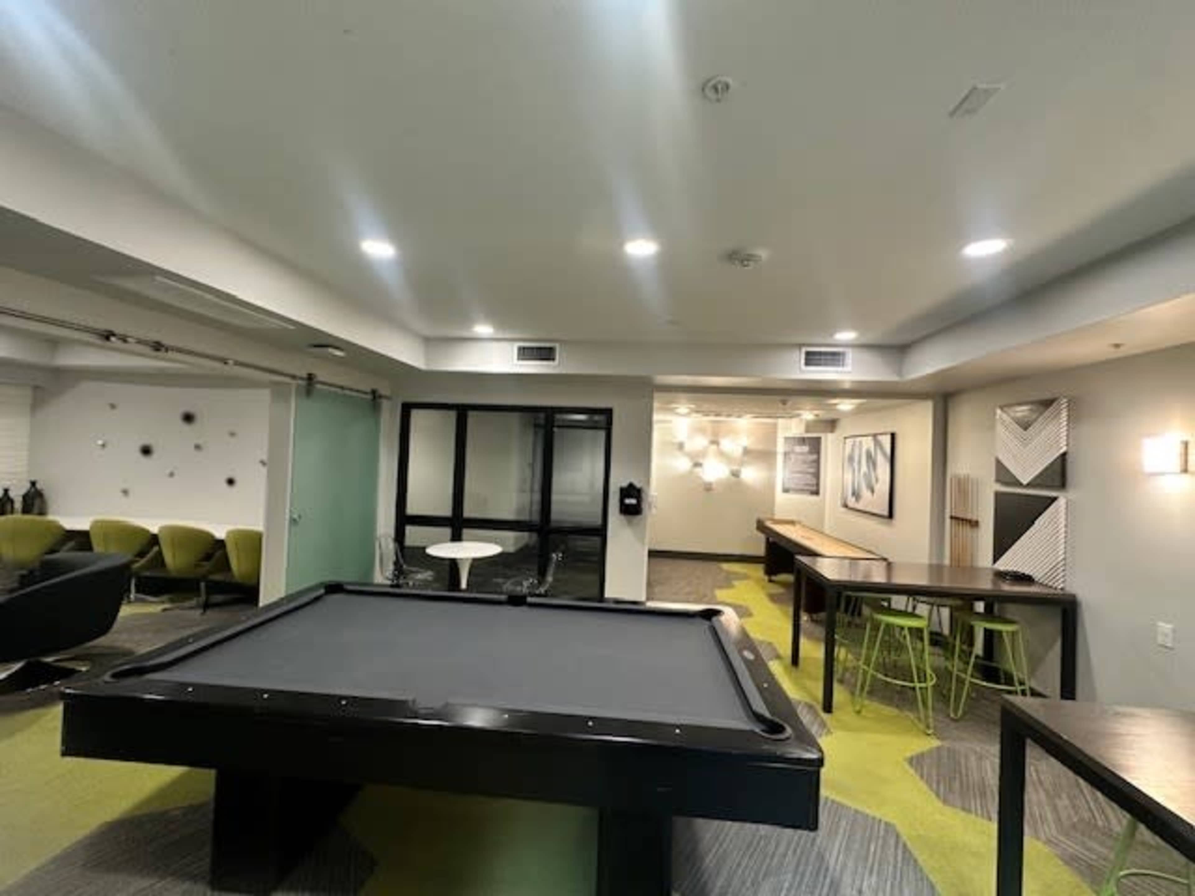 The image shows a modern common area with green carpet, a pool table, seating areas, and a lounge-style setup.