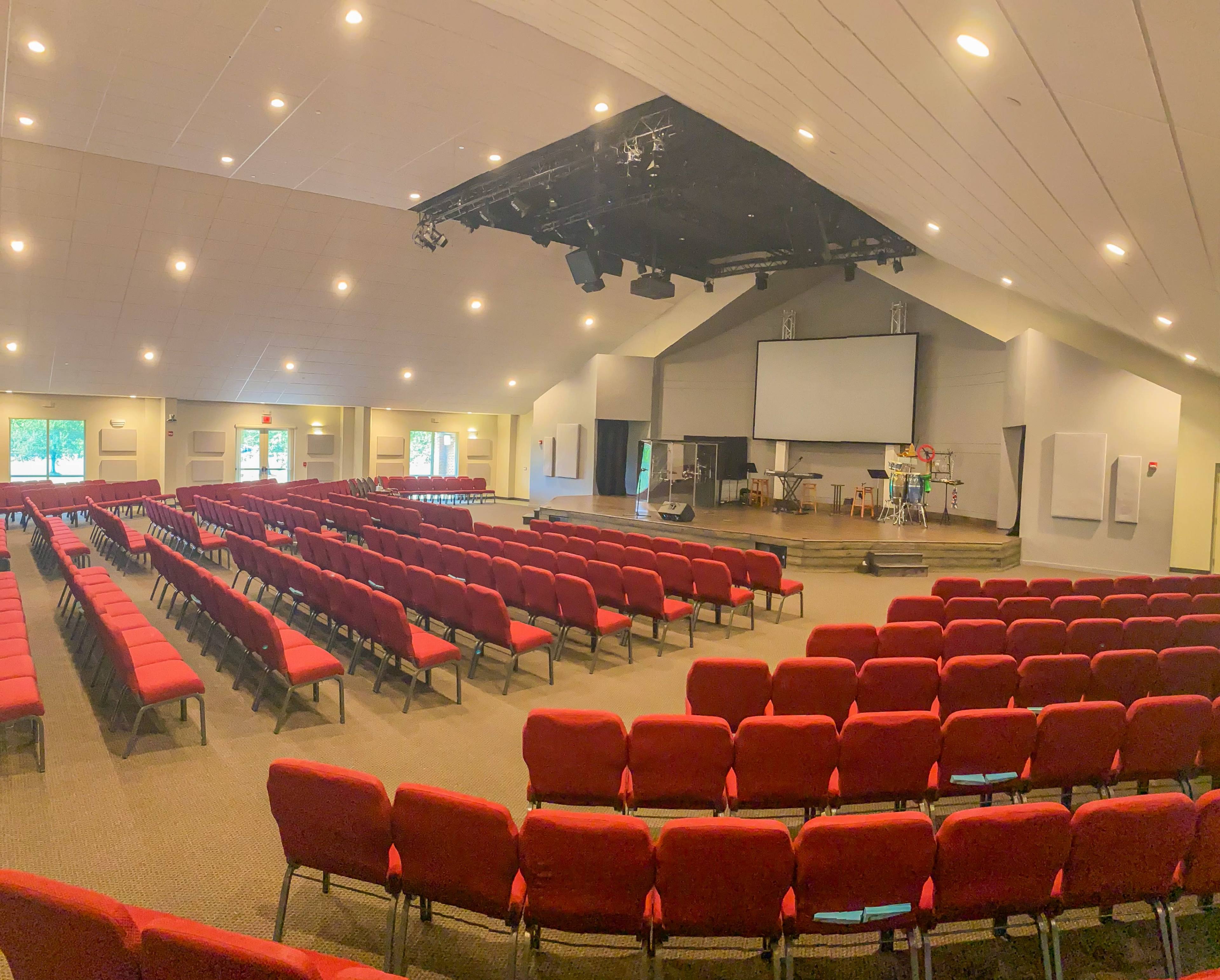 A large, empty auditorium with rows of red chairs facing a stage equipped with sound equipment and a projector screen.