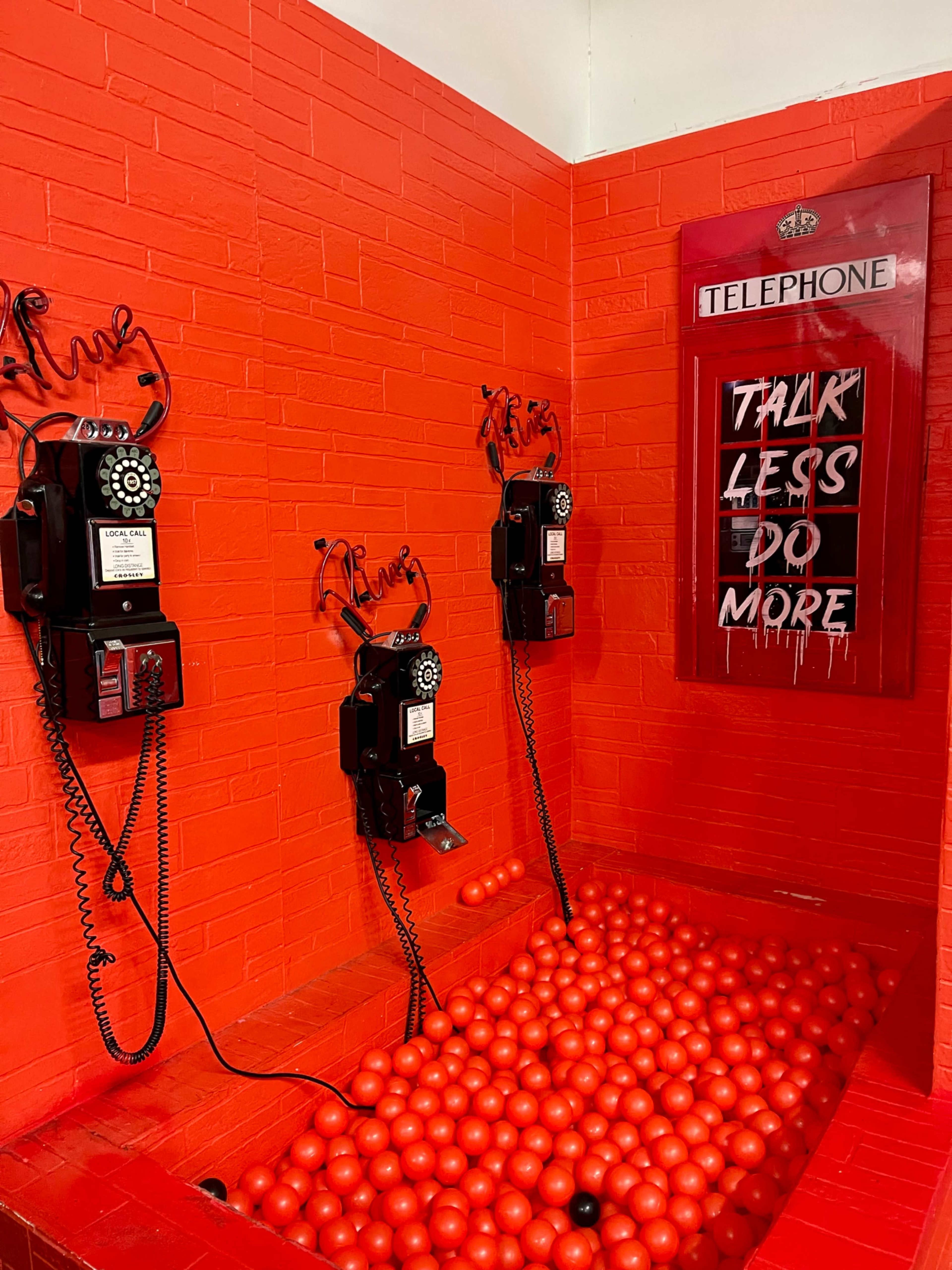 The image shows a bright red installation featuring three vintage black telephones mounted on a wall and a pool of red balls on the floor beneath them.