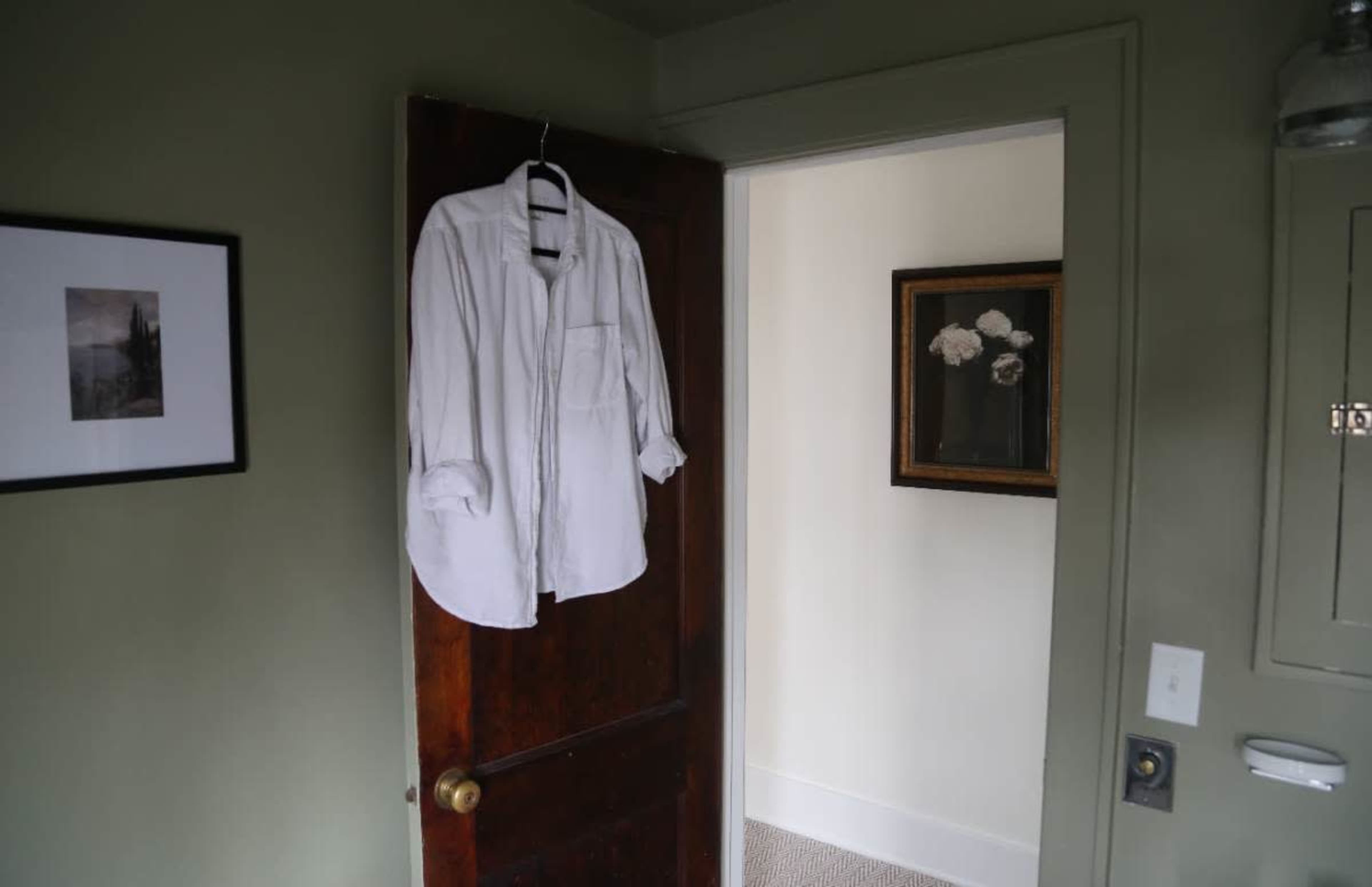A white shirt is hanging on the back of a wooden door, leading to a light-colored room with a framed painting on the wall.