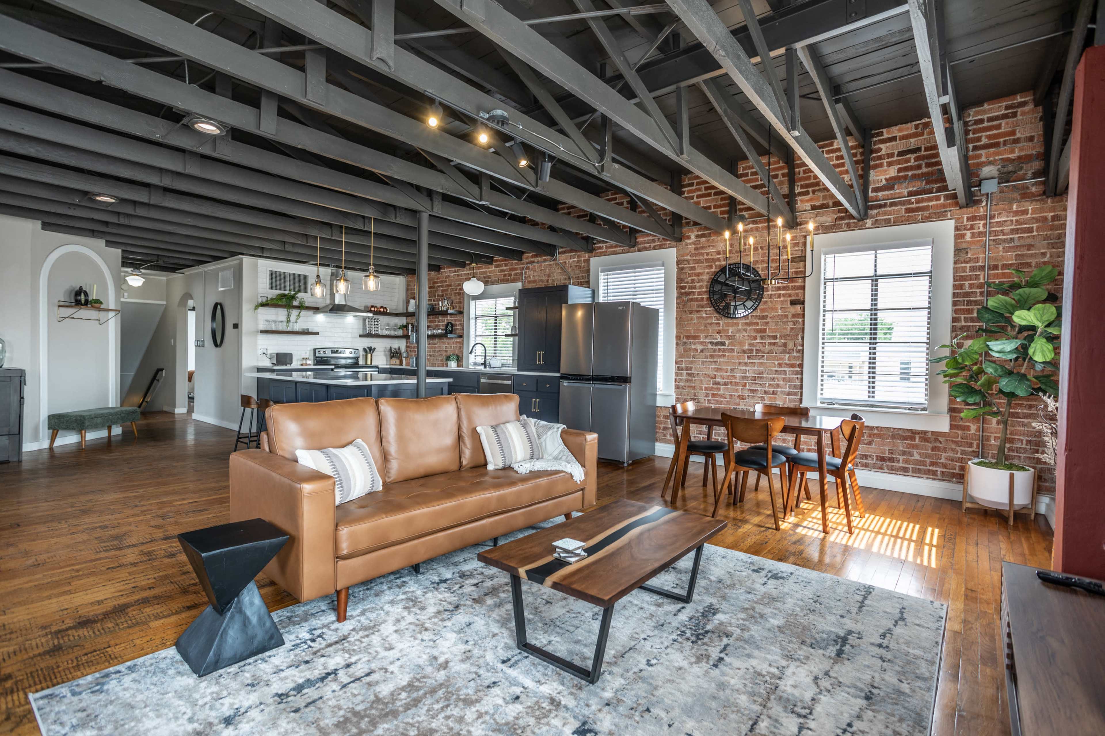 A modern loft-style living space with a leather sofa, a wooden coffee table, and a dining area featuring a round table and chairs, all set against exposed brick walls and dark beams.