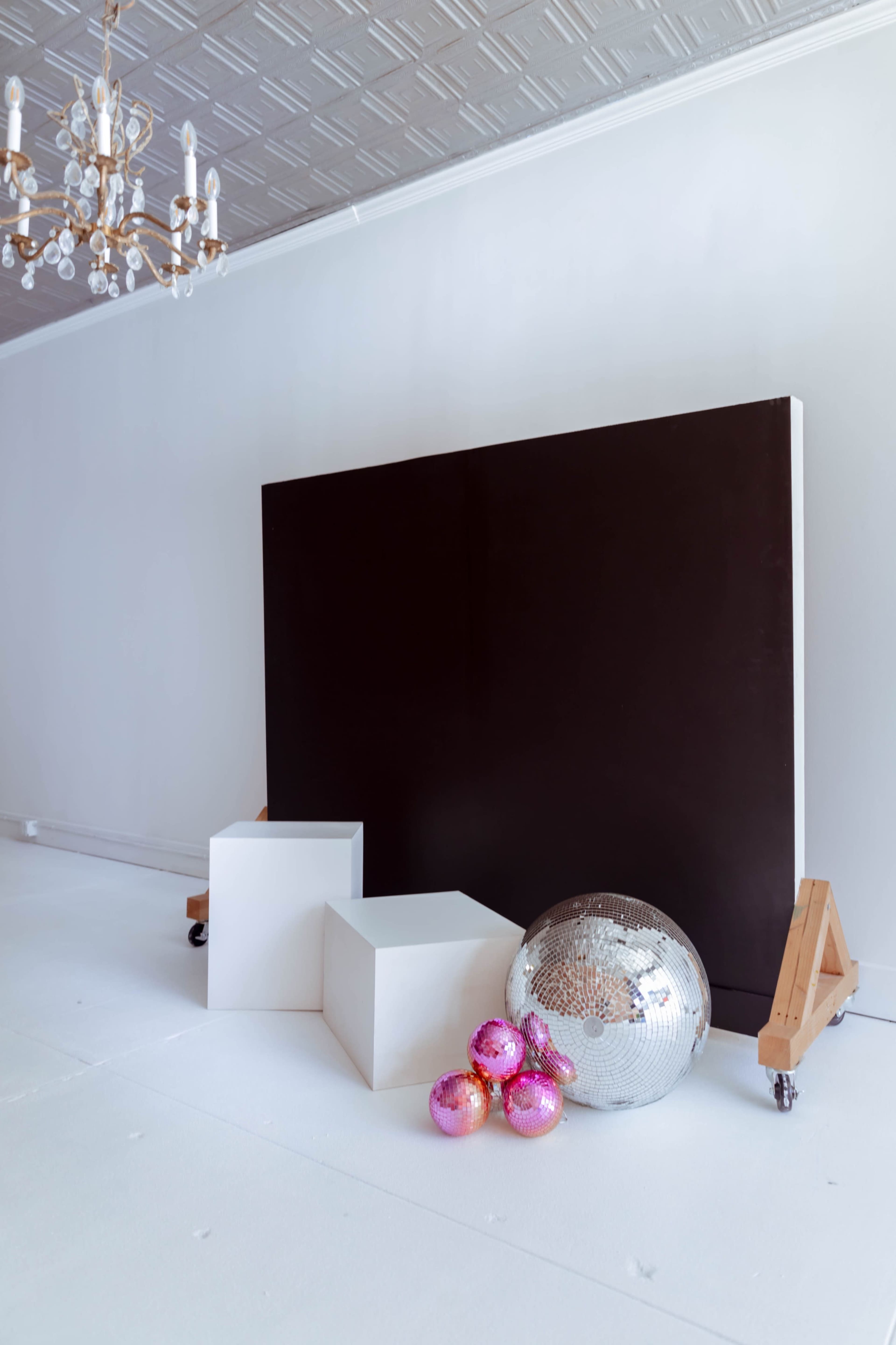 A modern photography studio features a large black backdrop, two white geometric pedestals, and colorful disco balls on the floor.