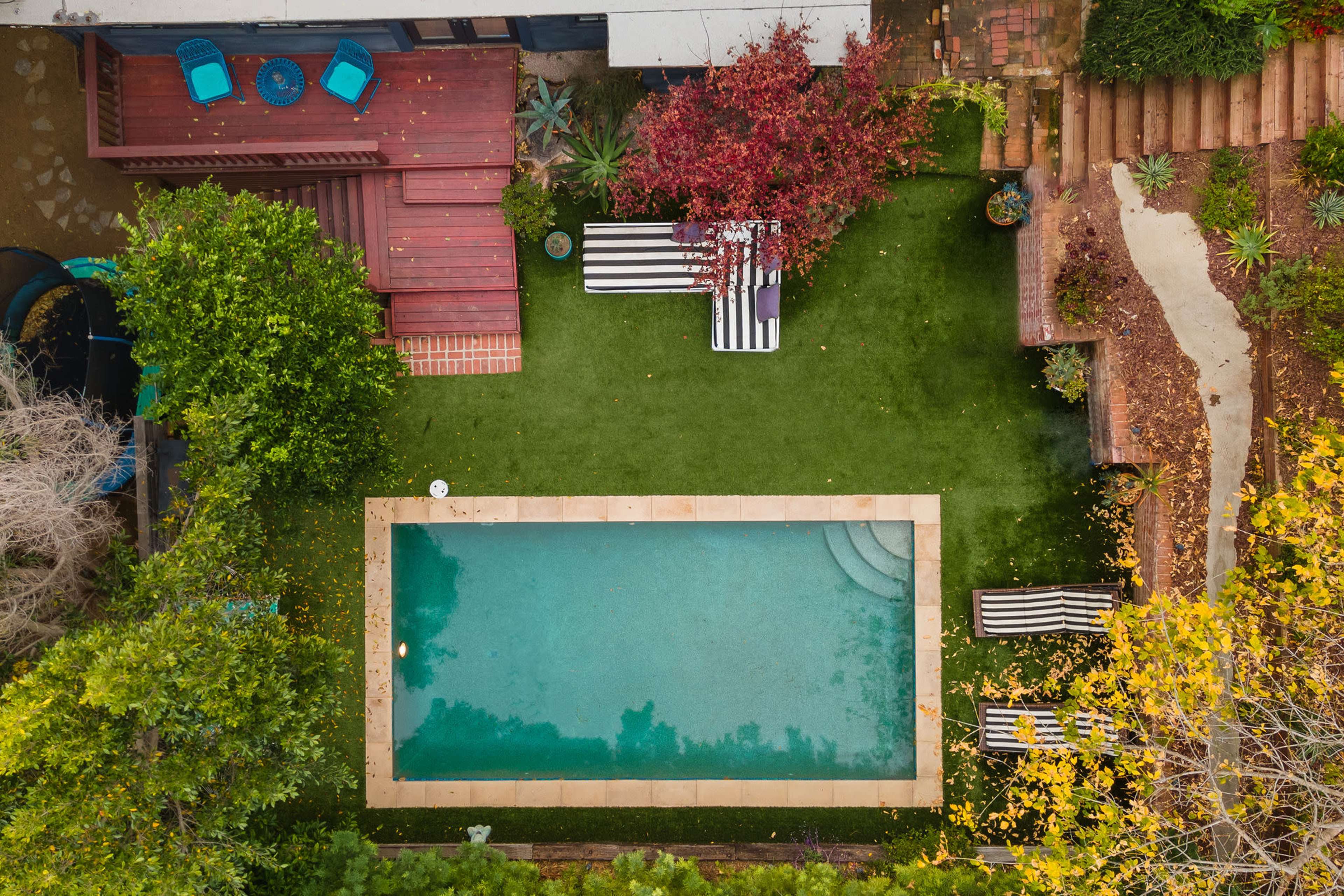 An aerial view of a backyard features a small swimming pool surrounded by green grass, striped lounge chairs, and wooden decking.